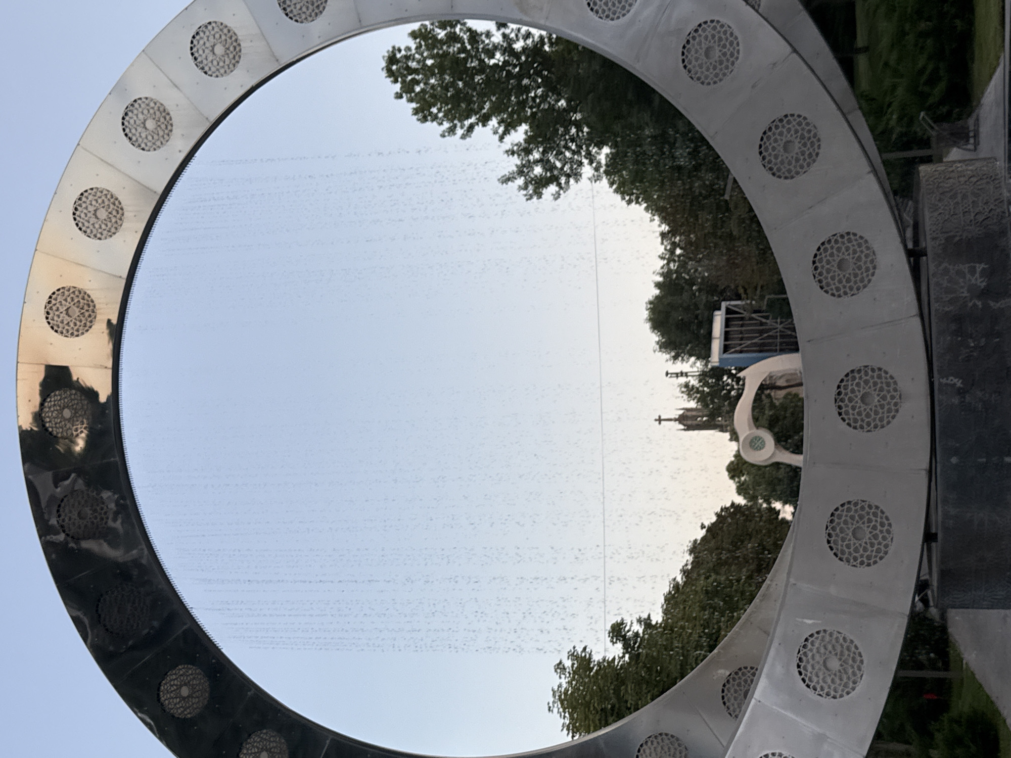 This image captures a close-up of a large, circular metal sculpture with decorative circular patterns on its surface. The sculpture appears to be a water feature, as a fine mist or curtain of water is visible falling within the circle, obscuring the background. Reflected in the water are trees and a clear blue sky. In the distance, through the water, a church spire with a cross is visible, along with a glimpse of green trees. The sculpture is situated outdoors, possibly in a park or public space within the city of Tashkent, Uzbekistan. The overall impression is one of public art and a tranquil, possibly scenic, environment.