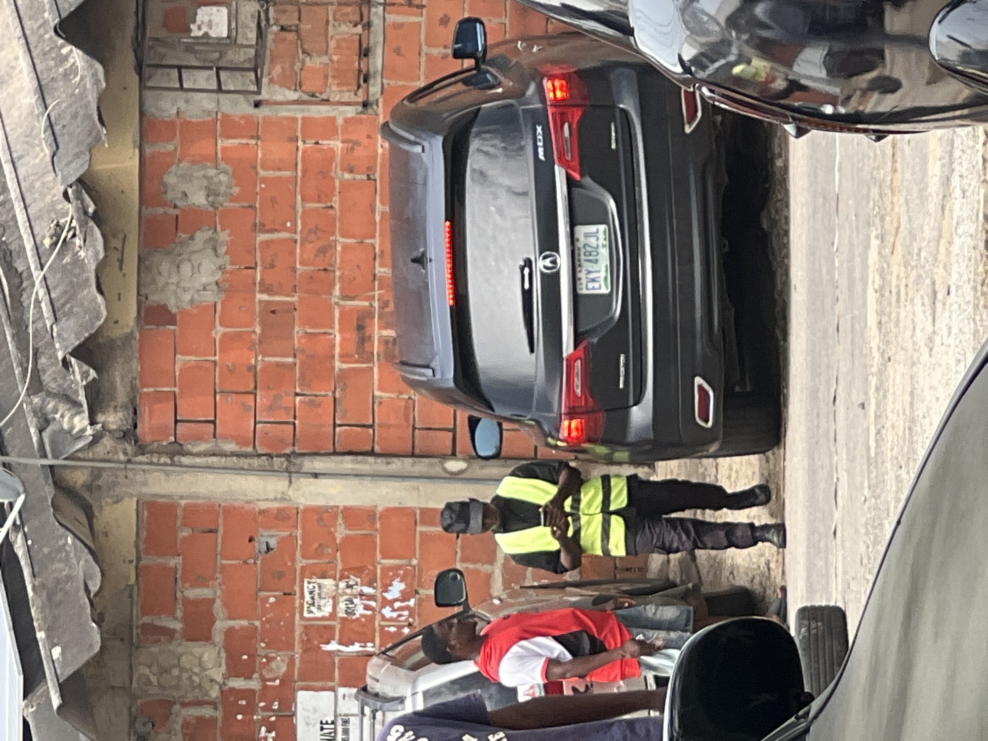 The image shows a street scene in Abuja, Nigeria, with an Acura MDX parked next to a brick wall. The license plate is visible and reads "EKY 482 JL." A man wearing a yellow and black reflective vest stands near the rear of the vehicle. Another person, wearing a red and white shirt, is lying on a car hood. The street appears narrow, with other vehicles visible on either side. The environment includes a corrugated metal roof overhang on the left and a partially obscured building with text signage.