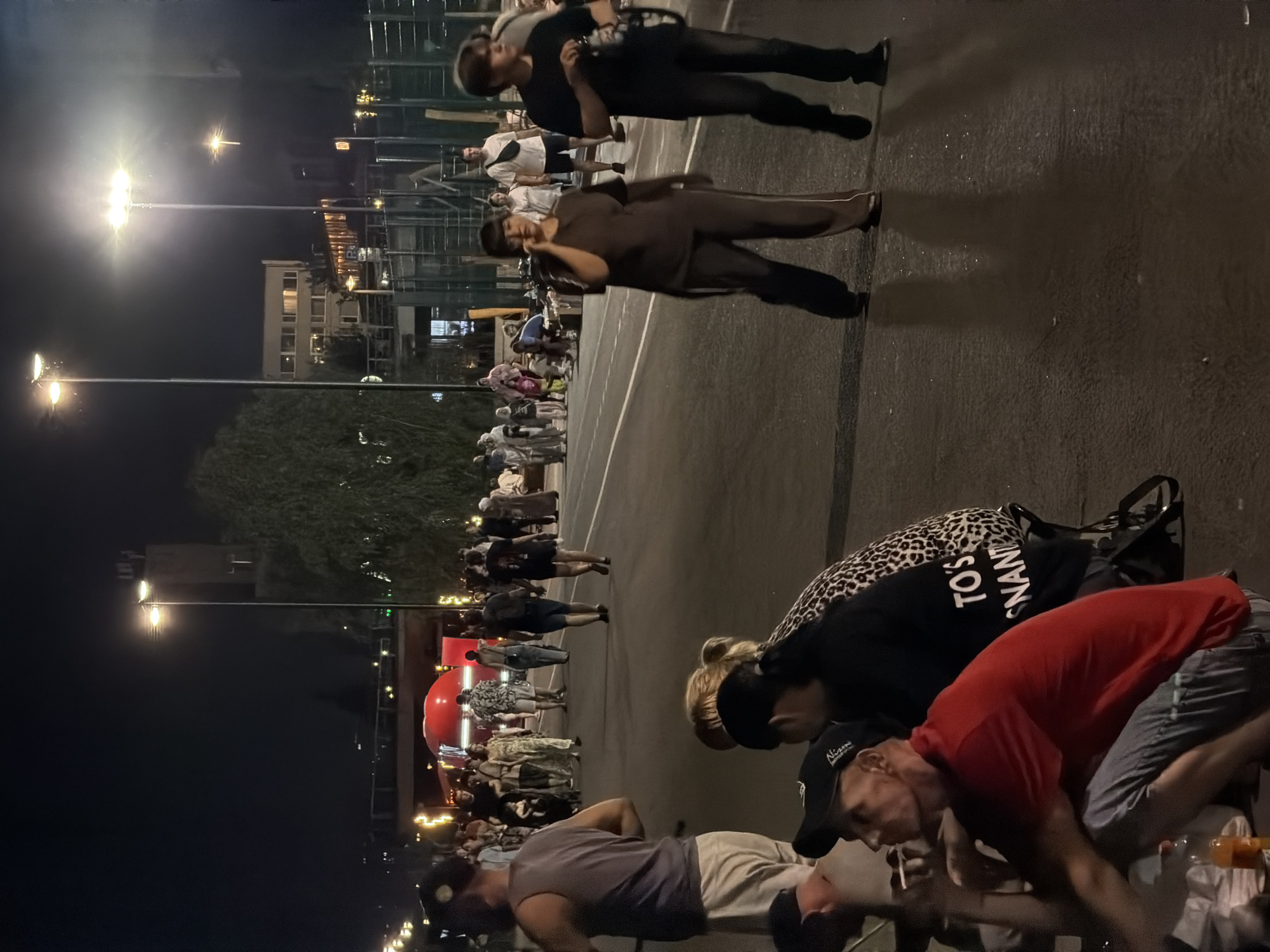 The image depicts a nighttime scene in a city environment, possibly Saratov, Russia, given the reference. A group of people are gathered on a paved area, which appears to be a street or walkway, illuminated by streetlights. In the foreground, several individuals are seated or squatting. One man in a red t-shirt and blue jeans is squatting, looking to his left, with a cigarette in his hand and an orange bottle nearby. To his right, a person wearing a black shirt with a leopard print pattern is seated, their face obscured. Further back, a woman in a brown tracksuit is walking and speaking on a mobile phone, while a young man in a black t-shirt walks beside her. In the background, a crowd of people are walking and standing, with a large red vehicle that resembles a food truck or bus visible. Buildings and trees are also present in the background, with several streetlights casting bright illumination. The overall atmosphere suggests a public gathering or event taking place at night.