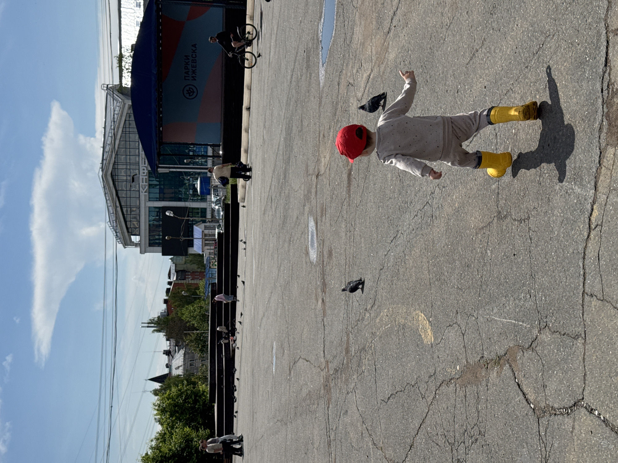 A child wearing a red hat and yellow boots is standing on a cracked paved surface in front of a building with a blue awning in Izhevsk, Russia. The building has a sign in Russian that says "PARKI IZHEVSKA." There are several pigeons on the ground near the child. A person on a bicycle is in the background. The sky is blue with some clouds. Trees are visible in the background, as well as other buildings and structures.