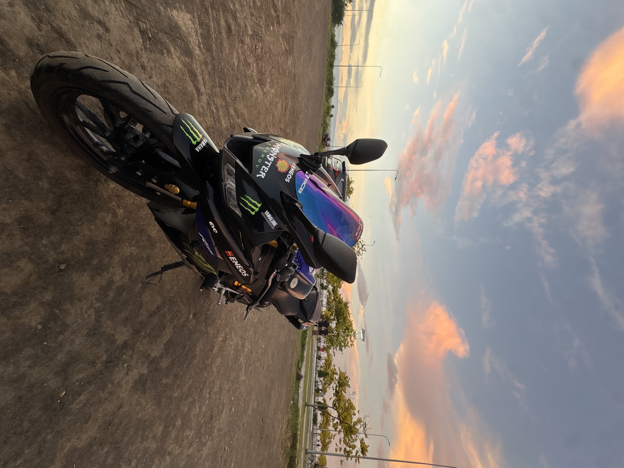 A black Yamaha R15 motorcycle is parked on a dirt lot with a coastal promenade in the background. The motorcycle is adorned with racing decals, including "Monster Energy" and "ENEOS." The sky is filled with colorful sunset clouds, tinged with pink and orange. In the background, trees line a paved walkway where a few people and a bicycle are visible. The scene is set during the golden hour. It is highly probable that this location is along the coastal road of Davao City, Philippines, given the typical scenery and the presence of palm trees.