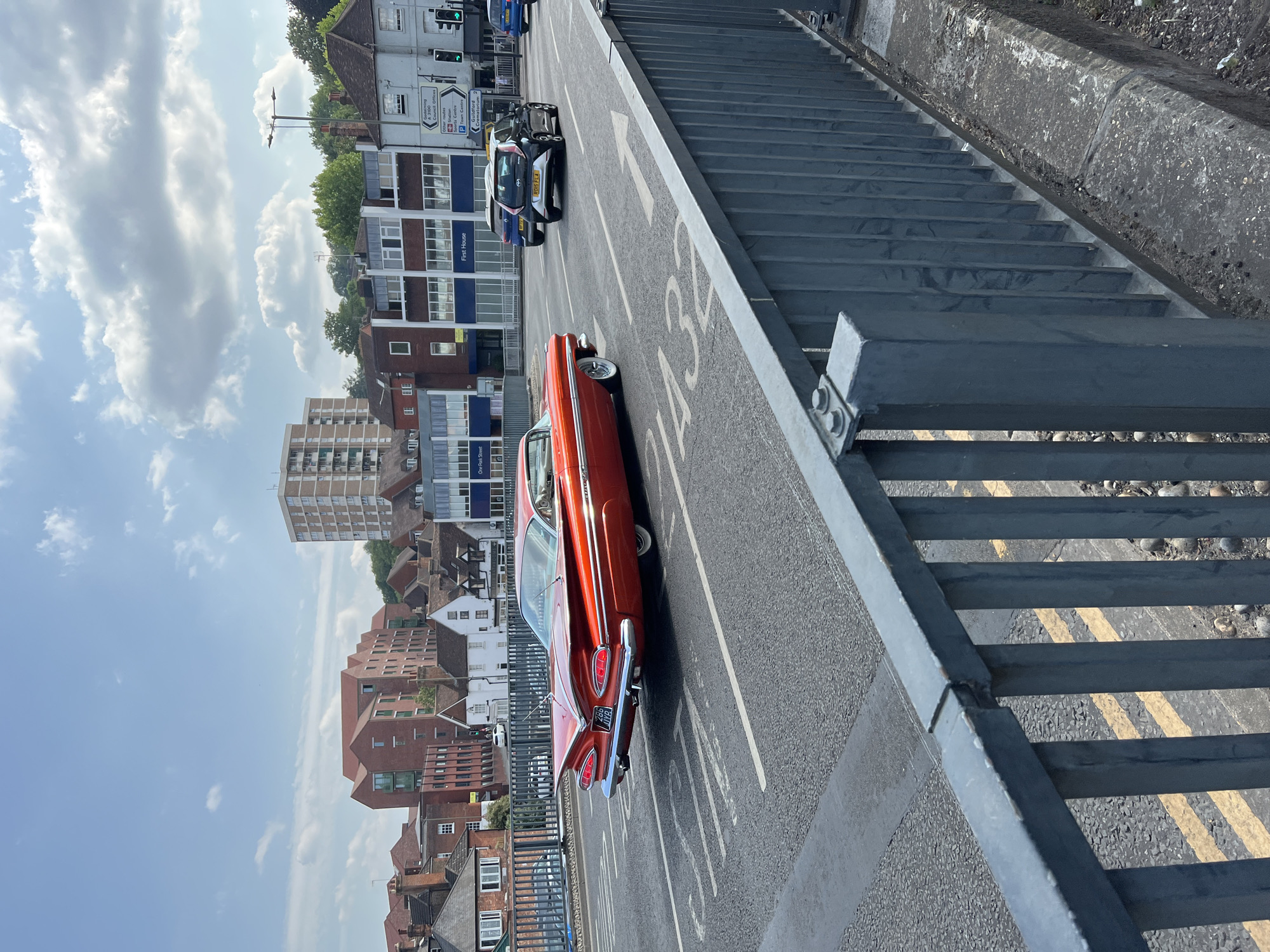 An orange vintage Chevrolet Impala is driving across a bridge in Guildford, United Kingdom. The car is viewed from the rear and is in the left lane of traffic. The bridge has a metal railing on the right side and is lined with double yellow lines. In the background, there are several buildings, including residential and commercial properties, and trees on a hillside. The sky is blue with scattered clouds. A white hatchback car and a blue minivan are also visible on the bridge, further ahead of the Impala.