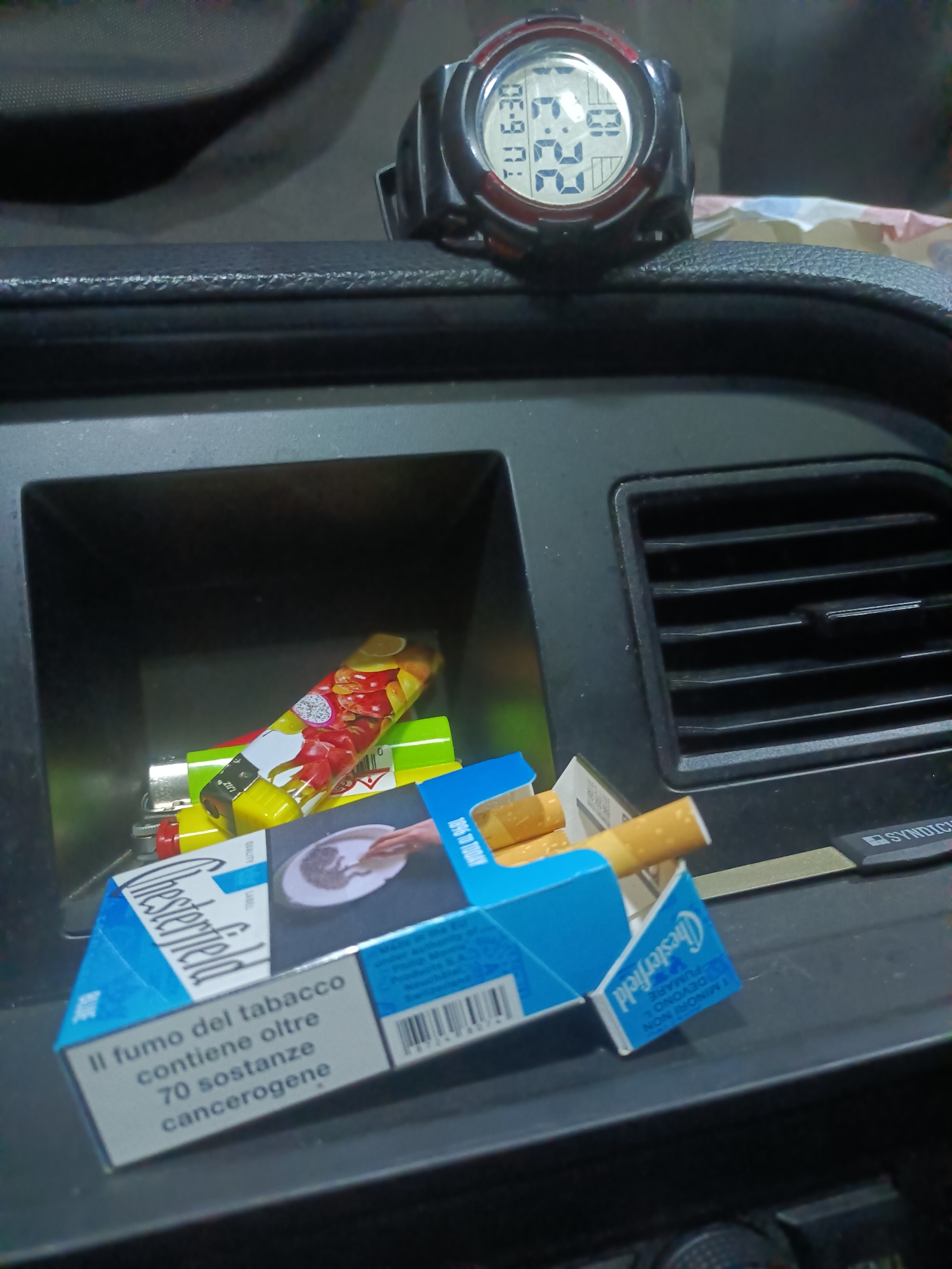 The image is a close-up, high-angle shot of a car's dashboard. In the foreground, a pack of Chesterfield cigarettes is open, with several cigarettes visible. The pack is blue and white and displays Italian text regarding the health risks of smoking. To the left of the cigarette pack, several lighters are visible inside a compartment, one with a colorful fruit design. Behind the cigarette pack and lighters, a digital watch is perched on the dashboard. The watch face displays the time "6:30" and the day "TU". The dashboard itself appears to be made of black plastic. A black fabric object is visible in the upper portion of the image, possibly a car seat or a bag. An air vent is visible to the right of the cigarette pack. The scene suggests a person has placed these items on the dashboard of a vehicle. The location is a car in Bagno di Romagna, Italy.