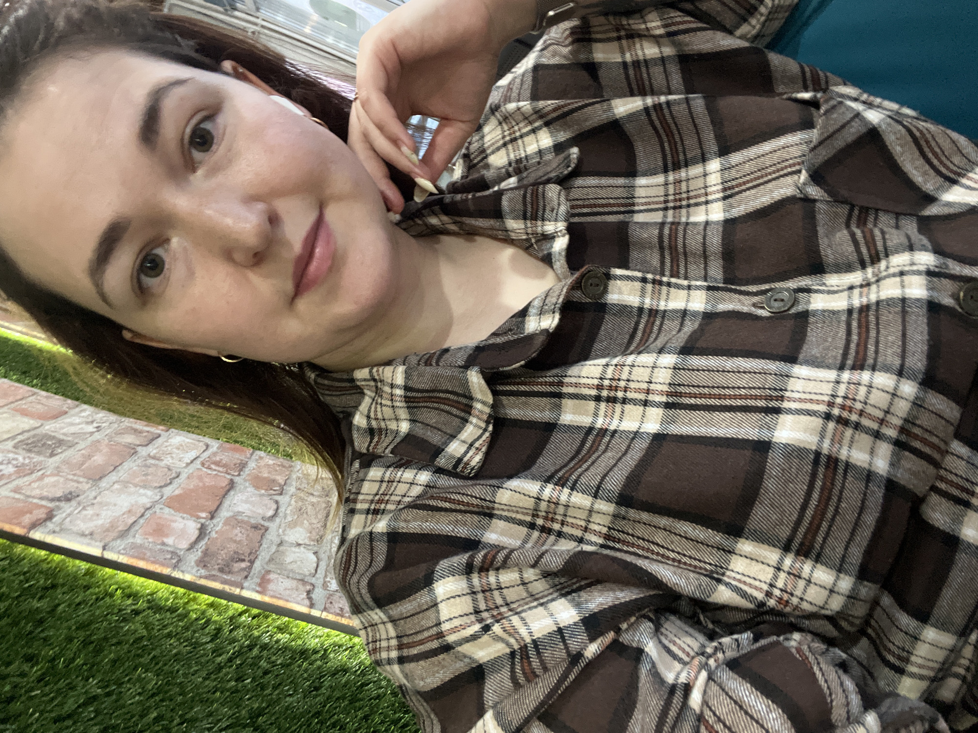 A young woman with dark hair and brown eyes is shown in a selfie. She is wearing a brown and cream plaid shirt and has on white earbuds and a small hoop earring. Her left hand is resting on her face and neck. The background shows a textured surface that resembles a brick path overlaid on artificial grass. The lighting suggests it is daytime. The image appears to have been taken indoors in a well-lit area, possibly a cafe or lounge with decorative elements.
