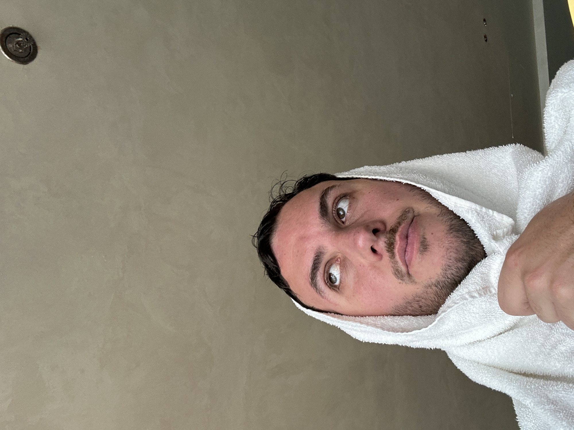 An image, originally captured in a portrait orientation but presented sideways, shows a man wrapped in a white towel. His head is positioned on the right side of the frame, with his face turned slightly to the left and his wide, hazel eyes looking upwards and to the left. He has dark, damp, curly hair and a short, dark beard or stubble. His expression appears curious, observant, or mildly surprised.
The man is covered from his head down to his shoulders by a thick, white terry cloth towel, suggesting he has recently bathed. The background is a plain wall in a light, warm grey or beige hue, featuring a subtle, stucco-like texture. In the upper left corner of the image (as if viewed upright), a small, circular metallic fixture, possibly a light or sprinkler head, is visible. Towards the upper right, two minor dark imperfections or small holes are present on the wall. The lighting is soft and even, indicative of an indoor setting. There is no visible text in the image. The context for this image is London, United Kingdom.