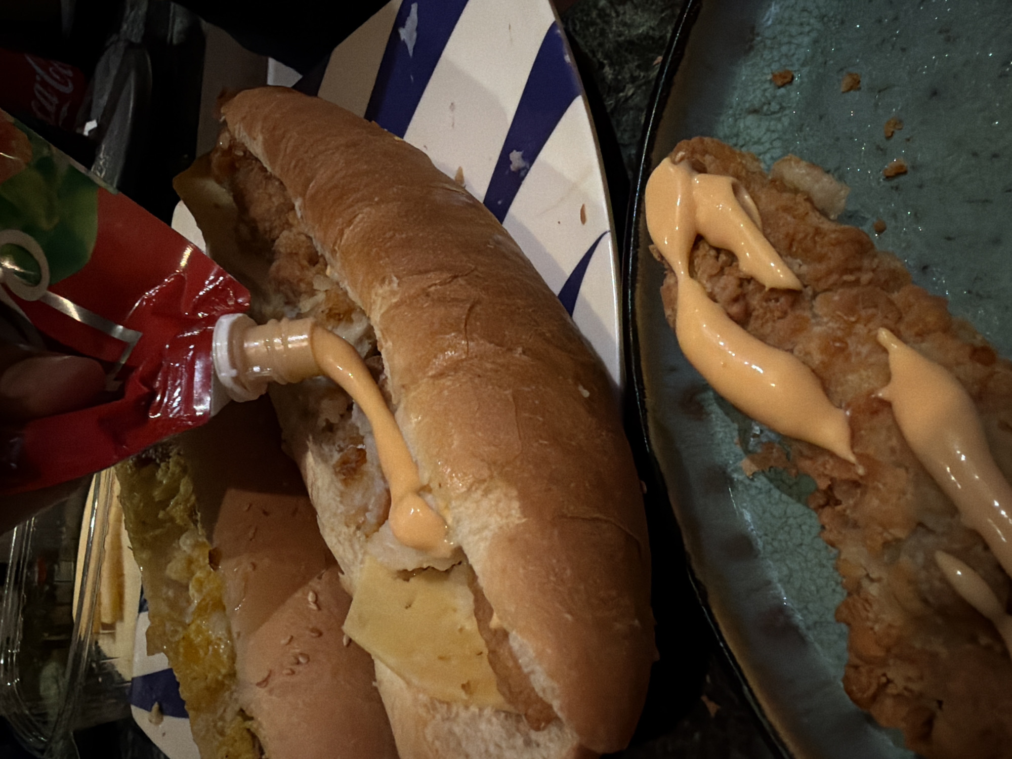 An overhead, medium shot captures a close-up view of two sandwiches, one in the foreground on a teal, textured plate and another in the background on a white plate with blue stripes. The sandwich in the foreground appears to be a piece of fried chicken on a bun, topped with a generous drizzle of orange-colored sauce. The sandwich in the background is a long roll filled with fried chicken and cheese, with more of the orange sauce being squeezed from a red pouch onto it. The scene is lit with warm, artificial light, highlighting the textures and colors of the food. The background is dark and out of focus, with a hint of a plastic container and possibly a Coca-Cola can visible. The image appears to be taken in a casual dining setting, possibly a restaurant or a home.