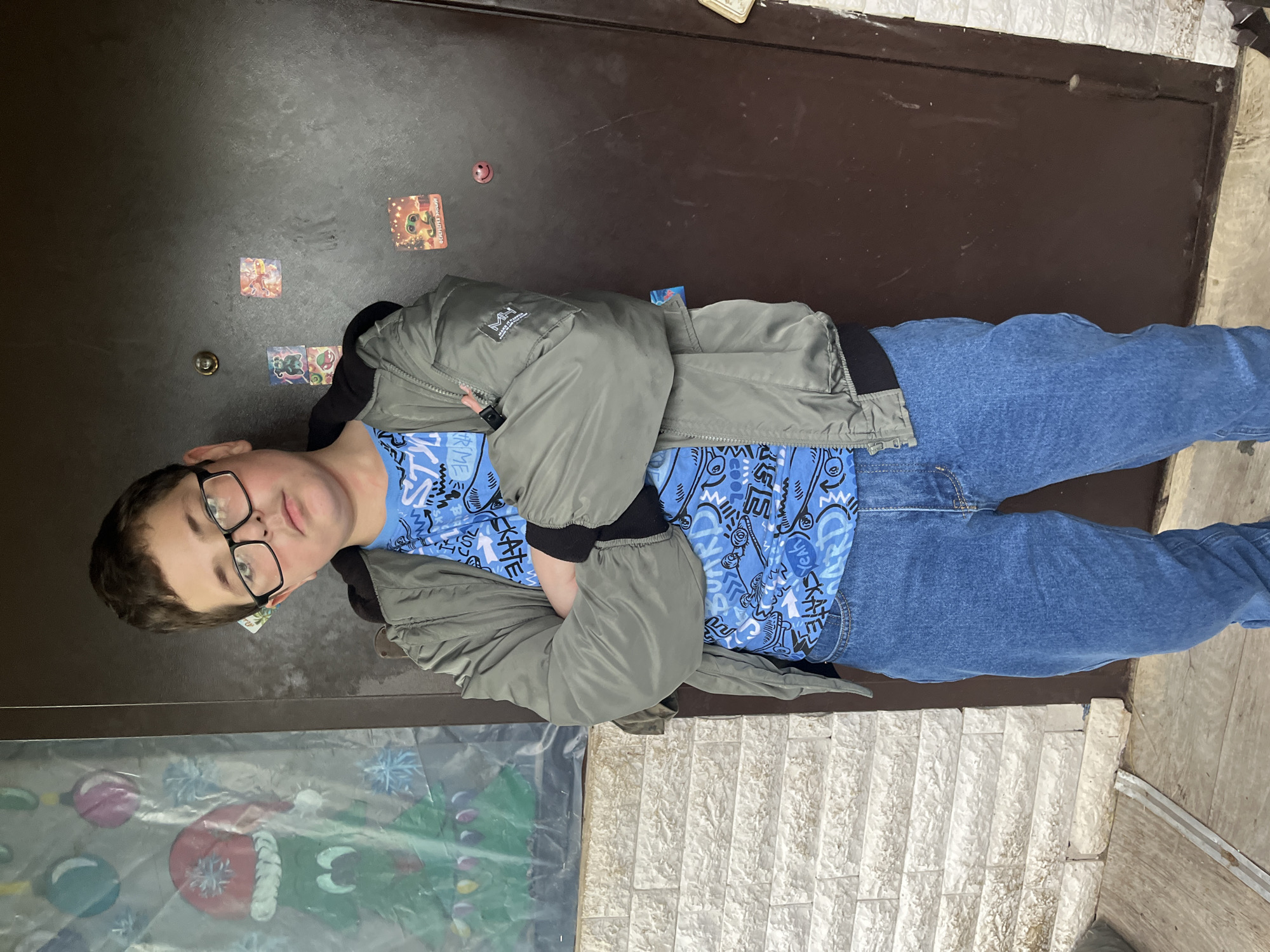 A young person stands indoors against a dark brown door. The person wears glasses, a light blue t-shirt with white and blue designs, a light gray jacket, and blue jeans. The arms are crossed. Stickers are on the door behind the person. The bottom of the image shows a painted or decorated surface. The location is Vesyoloye, Russia.