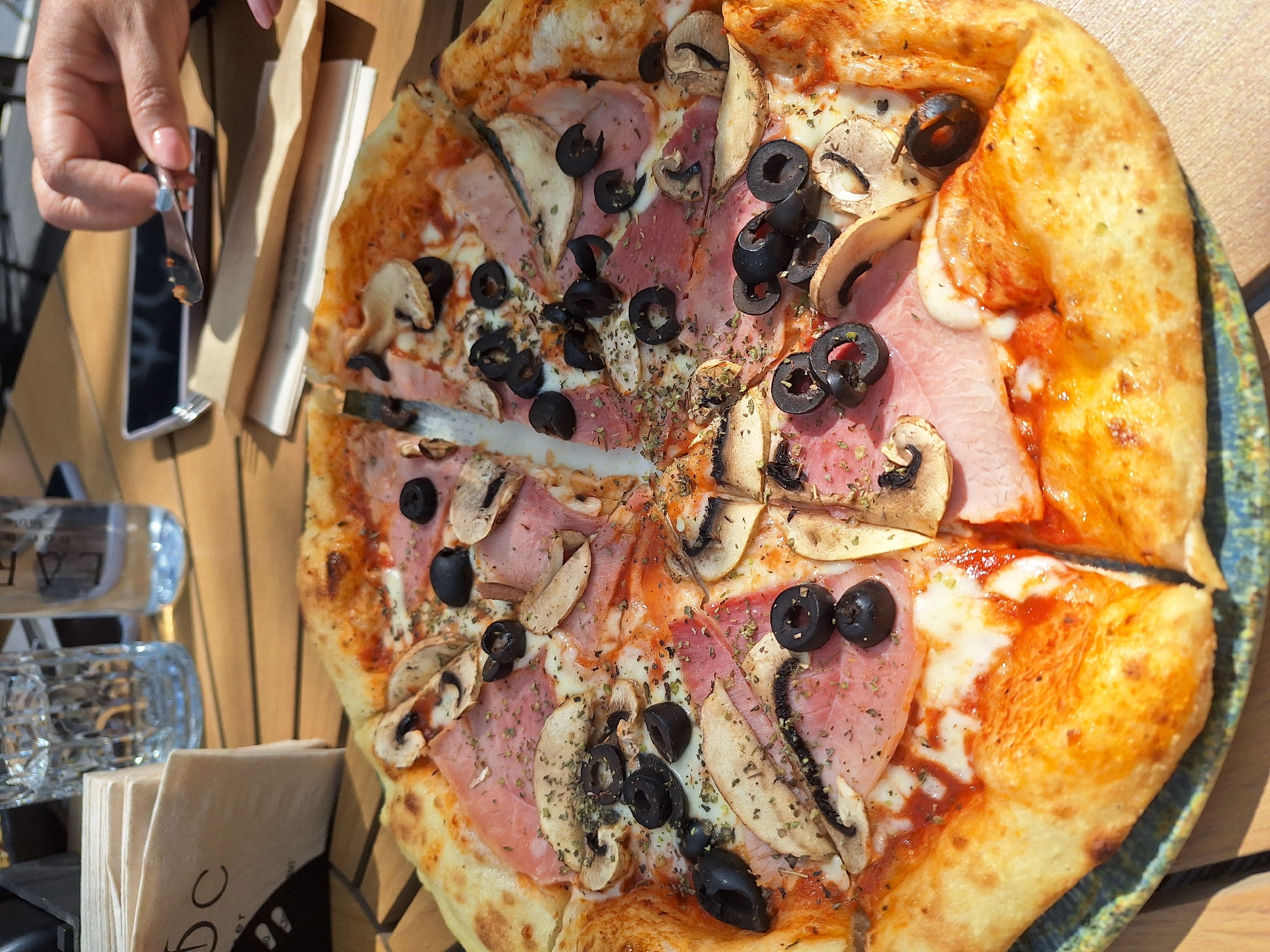 An overhead shot captures a vibrant pizza served on a wooden table, likely outdoors under bright daylight in Montana, Bulgaria. The central subject is a freshly baked pizza, generously topped with slices of ham, mushrooms, and numerous black olives. Dried herbs, possibly oregano, are sprinkled across the surface, enhancing its aroma. The crust is golden and puffed, with visible cheese and red tomato sauce peaking from underneath the toppings.
A person's hand, with light pink nail polish, is visible in the upper left, holding a fork with a small piece of food, suggesting they are in the process of eating a slice of the pizza. A smartphone, with its screen dark, rests beside a folded paper item that resembles a menu or bill holder.
Other objects on the table include a stack of brown napkins held in a metal dispenser, one of which partially shows the letter "C." A clear glass bottle, with the partial text "CAB" visible, and a clear glass ashtray are also present, indicating a casual dining setting, possibly a cafe or restaurant patio. The wooden table appears slatted, and strong shadows from the bright sunlight are cast across the scene, highlighting the sunny weather conditions. The pizza is already cut into slices, some of which appear to have been recently separated.