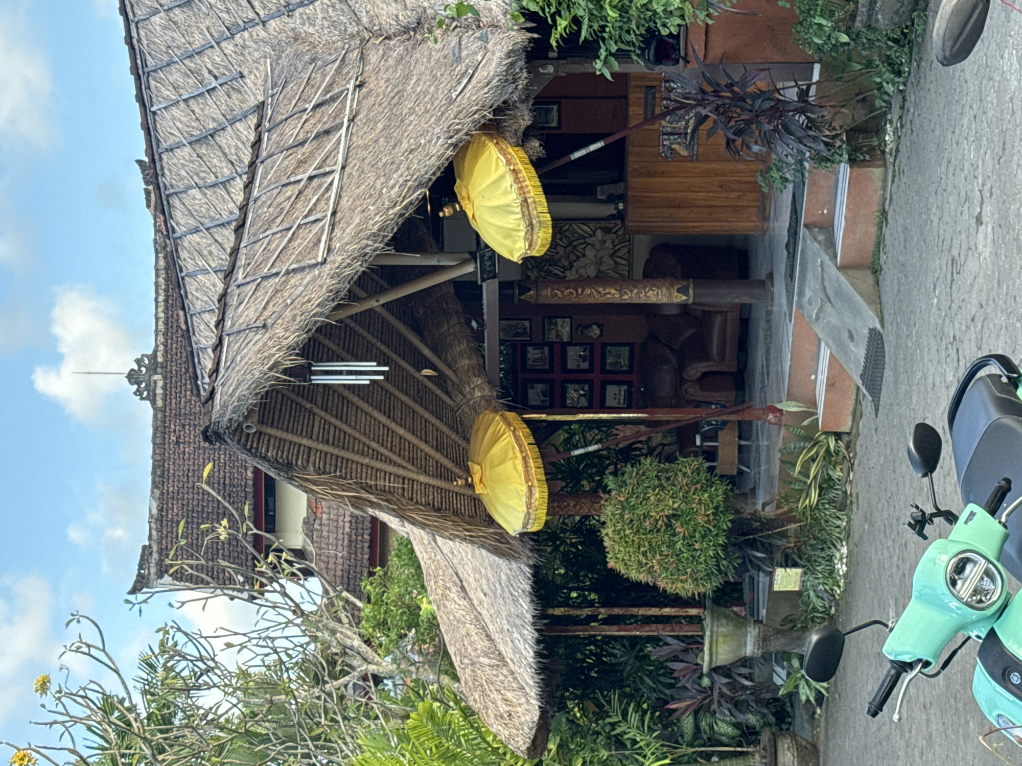 A daytime outdoor shot reveals a building with a thatched roof adorned with two yellow umbrellas. The structure is surrounded by lush greenery and a motorcycle is parked in the foreground. The building is located in Kuta, Indonesia.