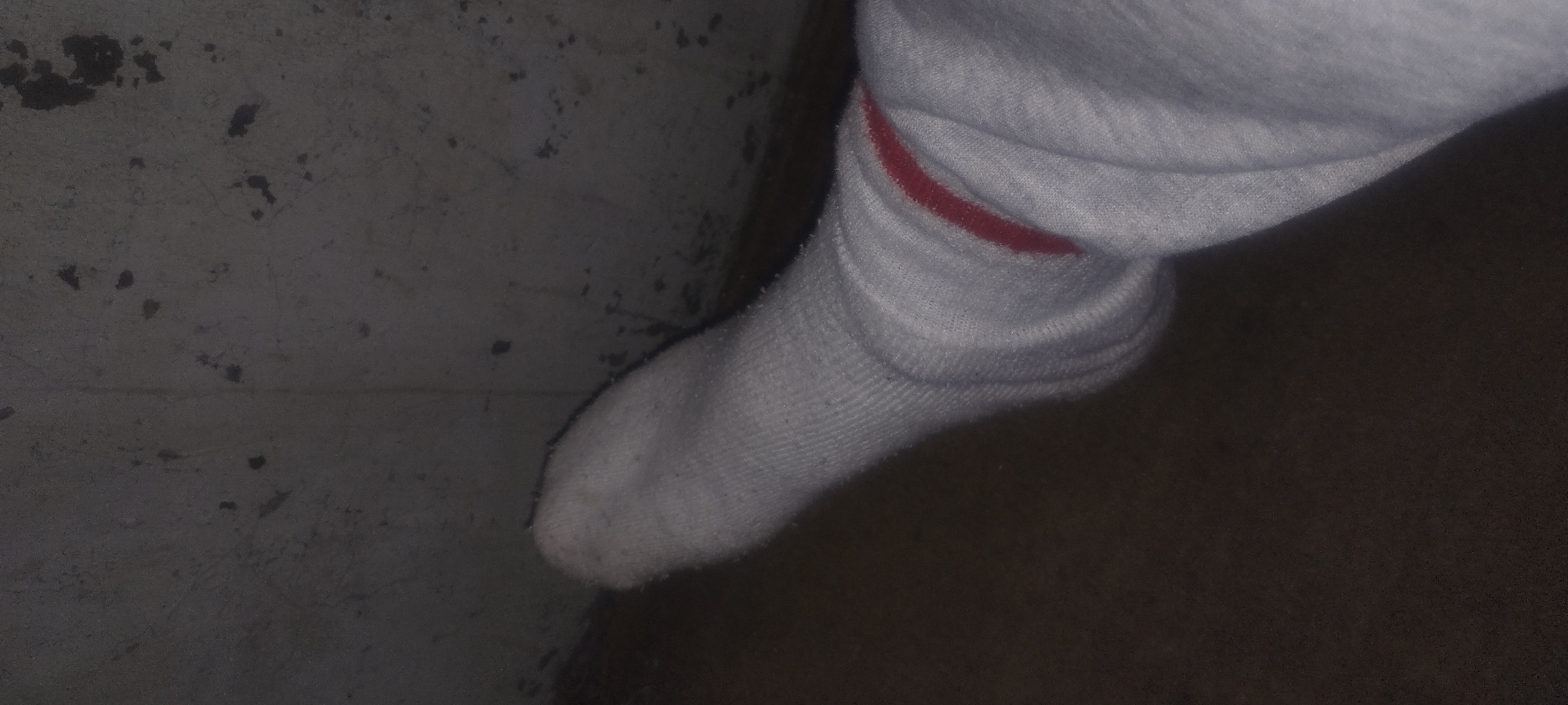 A close-up, low-angle shot shows a white sock with a red band around the ankle, worn by a person whose foot is angled towards the viewer. The sock is resting on a dark, textured surface, possibly a floor. Behind the foot, a rough, grey wall is visible, with dark splotches and textures. The lighting is dim, casting shadows and highlighting the fabric of the sock. The scene is likely indoors, given the wall texture and low lighting. There are no immediate indicators of the specific location within Zaporizhzhia, Ukraine, beyond it being an interior space with a concrete-like wall.