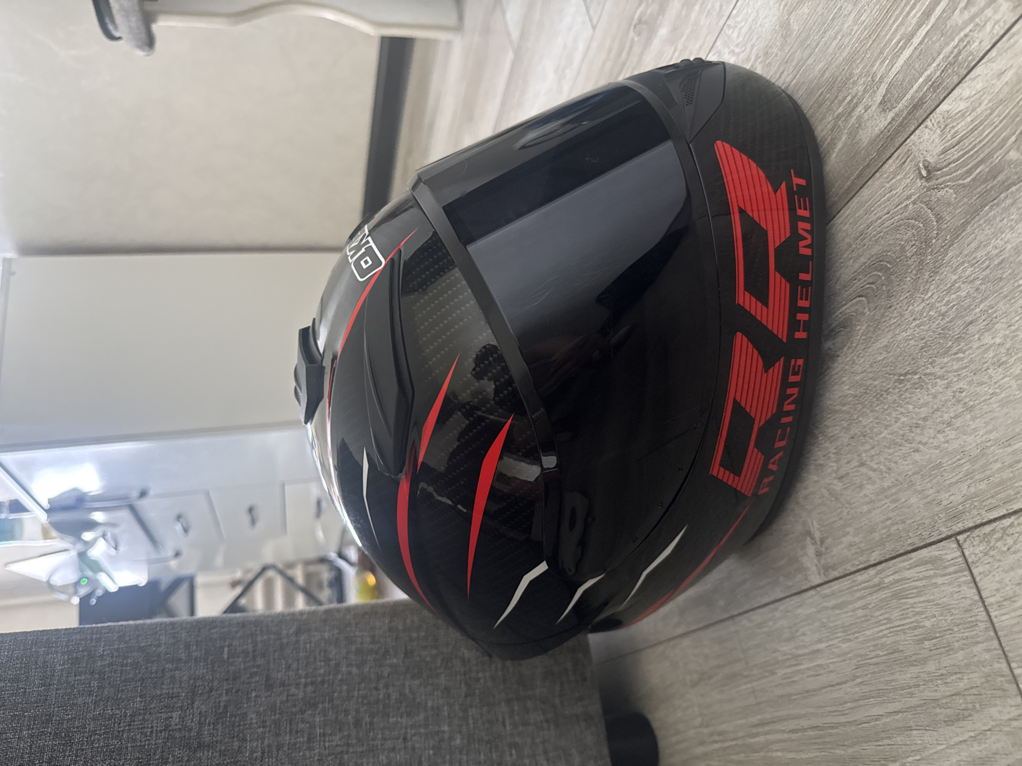 A black motorcycle helmet with red and white racing stripes and graphics rests on a light-colored wooden floor. The helmet has a dark tinted visor and the brand name "RR RACING HELMET" is visible in red lettering. In the background, a gray upholstered chair is partially visible on the left, and a white cabinet with drawers is on the right. The scene appears to be indoors, possibly in a residential setting.