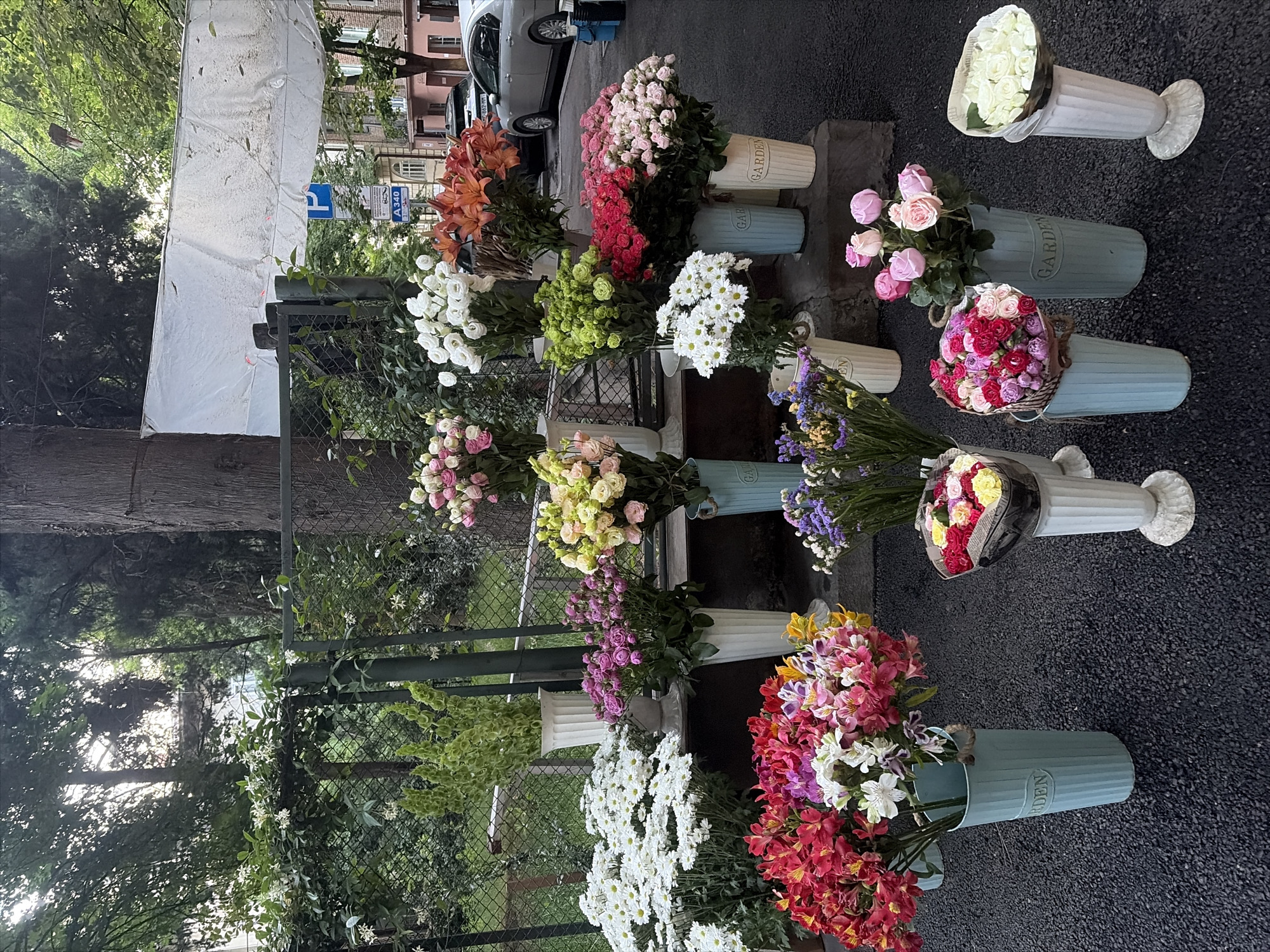 The image depicts a street-level outdoor flower stand in Tbilisi, Georgia, likely located in an urban neighborhood given the presence of parked cars and buildings in the background. A variety of floral arrangements are displayed in buckets and vases on the asphalt surface and on a low stone ledge. The flowers include white chrysanthemums, pink and white roses, red and pink alstroemeria, orange lilies, and smaller bouquets of mixed colors. Some of the buckets are light blue and have the word "GARDEN" embossed on them. In the background, a metal fence is visible, behind which is lush greenery and trees. A white tarp is stretched across the top of the scene, possibly to provide shade. A street sign with Georgian and Latin script is also visible. No people are present in the immediate vicinity of the flower stand.