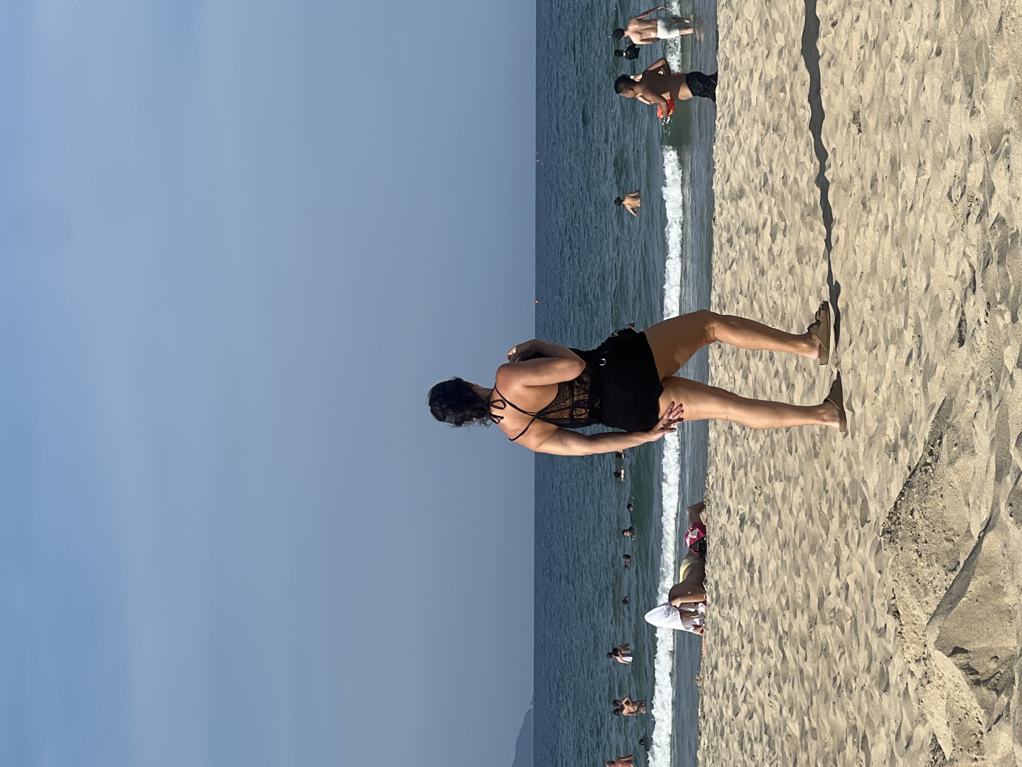 A group of people are enjoying the ocean and the beach on a clear day in Da Nang, Vietnam. In the foreground, a woman stands facing away from the camera, her dark lace-trimmed swimsuit and shorts visible. Her right hand is near her hip. To her left, a person lies on the sand, partially covered by a white towel, wearing a yellow and pink swimsuit. In the background, the blue ocean is populated with several individuals swimming and wading. A faint mountain range is visible on the horizon. The beach is sandy with some small mounds of sand in the foreground.