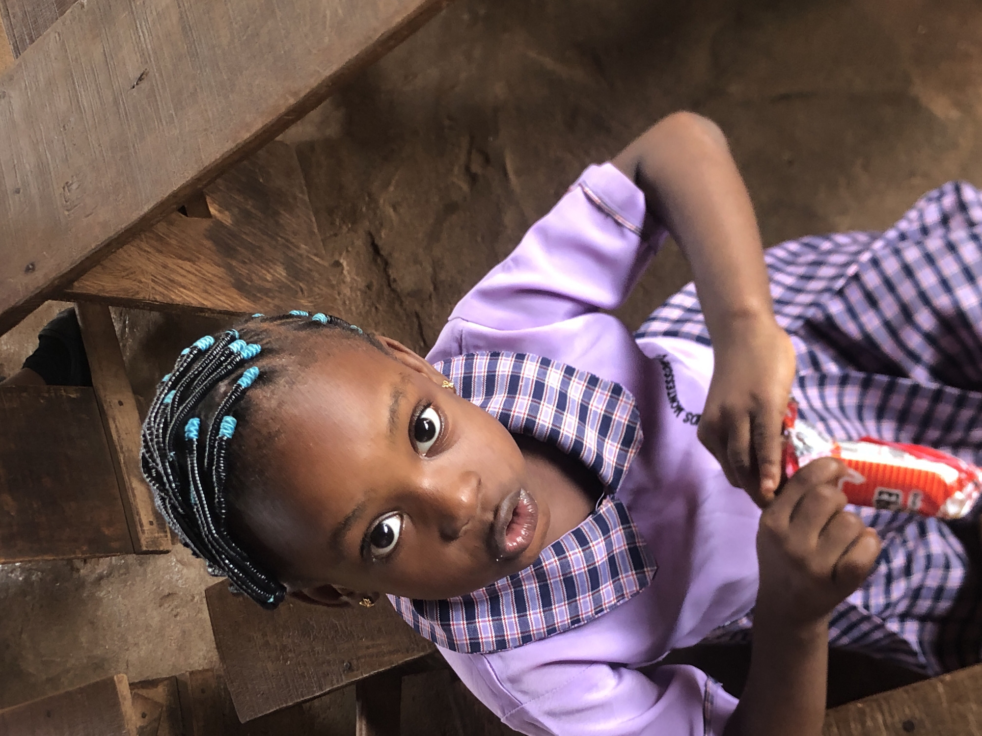 A young girl is looking up towards the camera while sitting on a wooden staircase in Ukwu-Abwa, Nigeria. She has dark skin, braids in her hair with blue beads, and is wearing a purple uniform with a plaid collar. She is holding a red and white packaged snack in her hands. The staircase is made of wooden planks, and the background behind the stairs appears to be a rough, earthen surface.