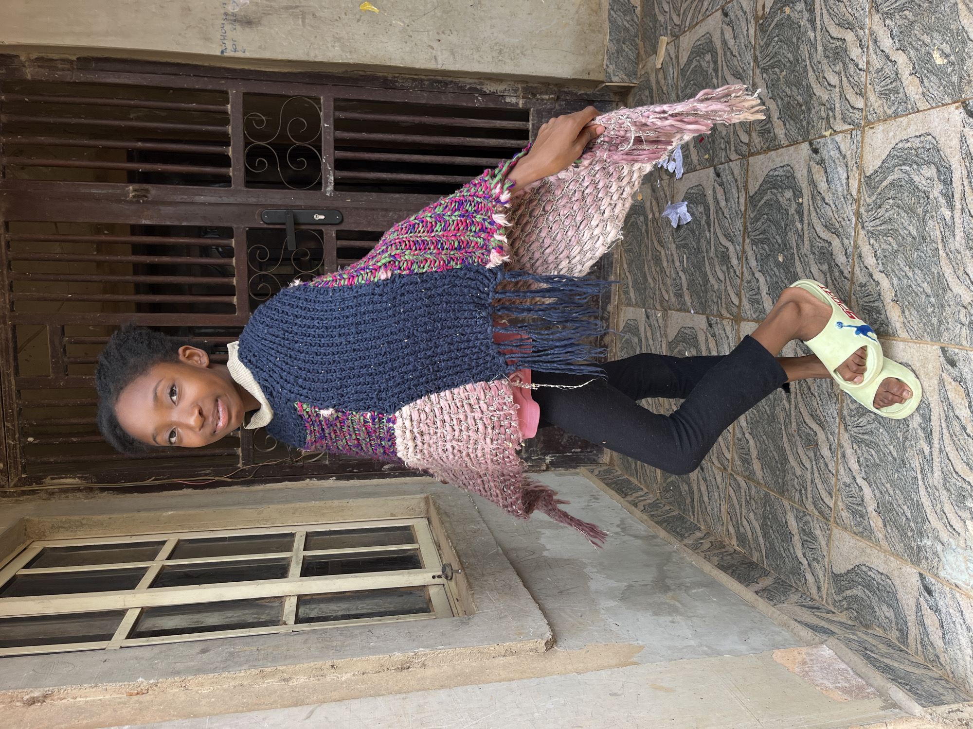 A young Black girl stands outside a building, her body angled slightly to her left. She wears a dark blue, crocheted poncho with a white collar, a pink shirt, black leggings, and light yellow slides. The poncho is adorned with colorful, textured panels and fringe. Her right hand is raised, grasping a portion of the poncho.
The background features a weathered, light-colored wall with a multi-paned window to the left. To the right, a dark brown metal gate is visible, partially obscuring the entrance to a building. The ground is tiled with a marbled pattern. The overall environment appears to be an urban setting.