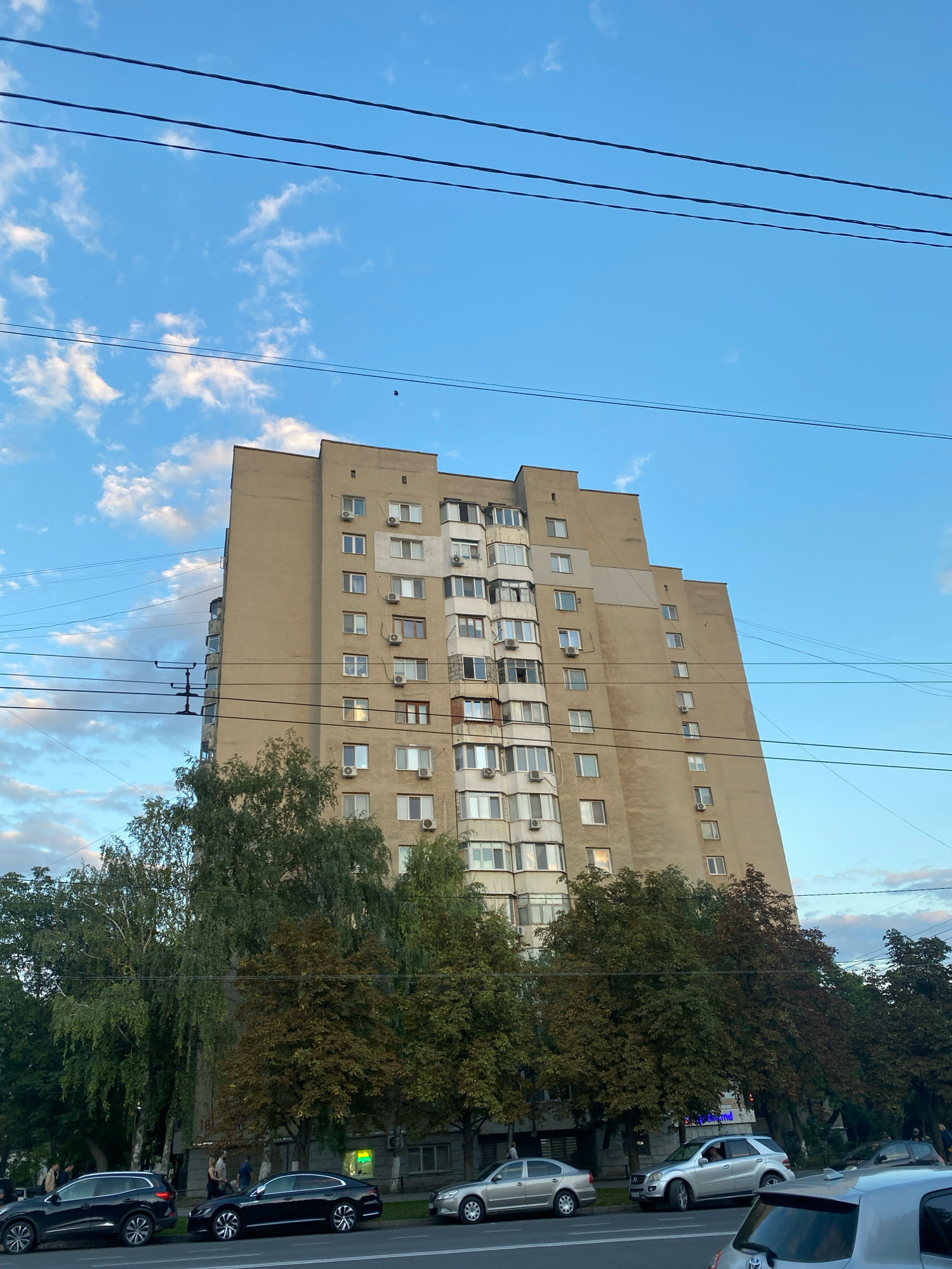 The image depicts a tall, beige apartment building in Chișinău, Moldova, likely in an urban residential area. The building has a stepped facade and numerous windows and air conditioning units. It is flanked by mature trees, some with green foliage and others with reddish-brown leaves, suggesting an autumn setting. Several cars are parked along the street in front of the building, including a black SUV, a black sedan, and a silver sedan. A few individuals are visible near the parked cars, some appearing to be walking or standing. Overhead, power lines crisscross the frame against a blue sky with scattered clouds. The scene conveys a typical city streetscape with residential architecture and natural elements.