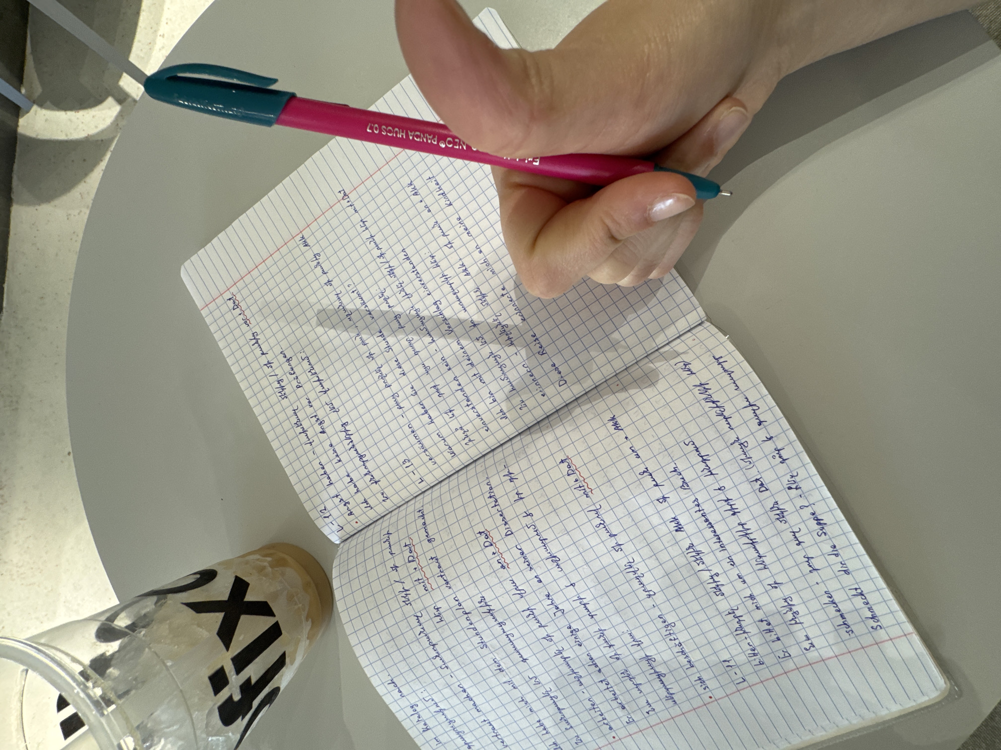 Here's a detailed description of the image:
**General Overview:**
The image is a close-up shot taken from an overhead perspective. It features a hand writing in a notebook, which sits on a light-colored surface, likely a table. Beside the notebook is a partially filled plastic cup, apparently containing an iced drink.
**People:**
Only a hand is visible, indicating the presence of a person writing. The hand is holding a pen. The hand has fair skin.
**Subjects:**
The subjects are the act of writing, a notebook, a pen, and a beverage.
**Objects:**
* **Notebook:** A spiral-bound notebook with grid paper (possibly a squared notebook). The notebook is open, with writing visible on both pages. Some text is underlined in red.
* **Pen:** A pink pen with a teal cap. There are inscriptions on the pen body, and the inscription NEO PANDA HUCS 0.7 can be seen.
* **Drink:** A clear plastic cup containing ice and a liquid. The cup has a black logo on the side.
* **Table:** A round, light-colored, possibly light grey table.
**Actions:**
The primary action is writing. The hand is holding the pen and is in the process of writing in the notebook.
**Interactions:**
* The hand interacts with the pen to write in the notebook.
* The notebook rests on the table.
* The cup sits beside the notebook on the table.
**Environment:**
The environment appears to be a cafe or workspace setting, with a plain light-colored table and a partial view of a chair in the background.
**Location:**
The image does not provide enough information to determine if the location is in Yerevan, Armenia.