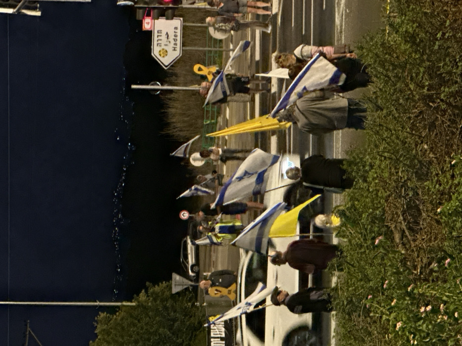 This image depicts a nighttime scene at a road intersection in Mishmar Hasharon, Israel. A group of approximately 15-20 individuals are gathered on the roadside and pavement. The sky is dark, with distant lights visible on the horizon in the background.
Numerous individuals are holding Israeli flags (blue Star of David on a white background with two blue stripes). Several yellow flags are also present, and multiple individuals are displaying or holding large yellow ribbon symbols, commonly associated with supporting hostages. One such yellow ribbon symbol is mounted on a stand on the far left. A partially visible banner with Hebrew text, including "ביחד" (together), is also present. Most people are casually dressed. One individual near the center-left wears a reflective yellow-green vest.
Vehicles are present on the asphalt road. A white car, possibly an SUV, is in motion in the center of the frame, its headlights illuminated. Another light-colored sedan is visible further back on the left. Traffic lights are visible on the right side of the intersection, displaying a red signal. A white directional sign points to "Hadera" (חדרה) in both Hebrew and English, featuring a yellow date palm icon. Additional road signs, including a yield sign and a no-entry sign, are visible on the left. The foreground features green foliage and shrubs, partially obscuring the lower parts of some individuals. Taller, drier vegetation lines the right side of the road. Artificial lighting from streetlights illuminates the scene.