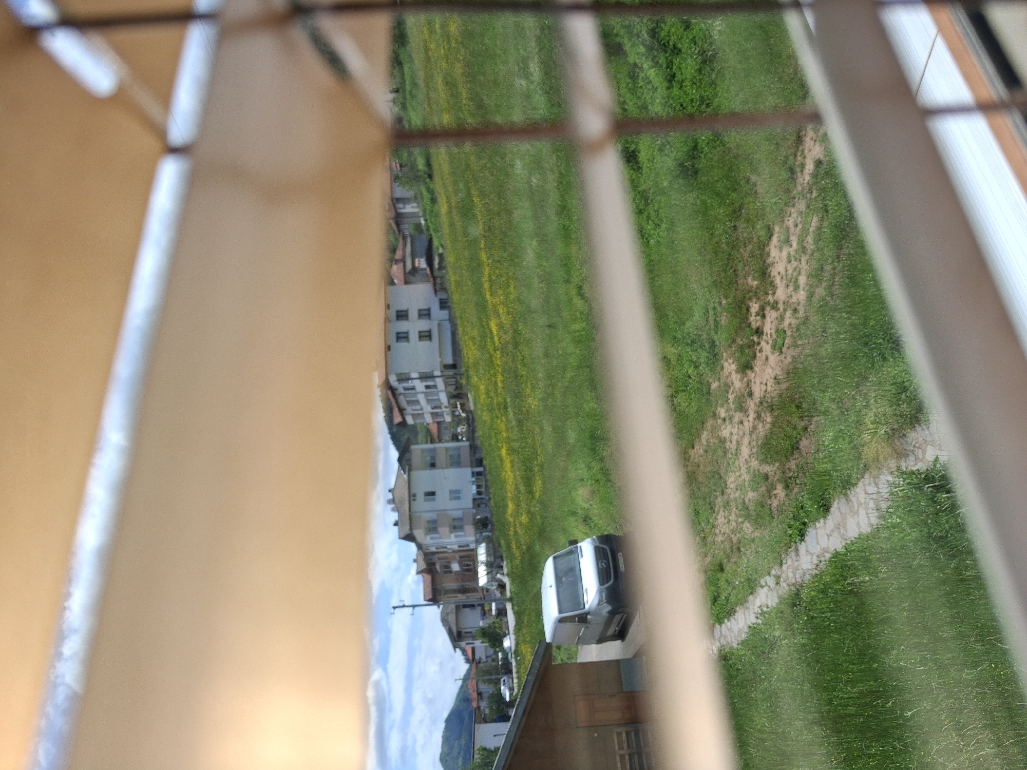 A view of a landscape with buildings, a van, and grassy areas, seen through the slats of a window covering. The buildings appear residential, arranged along a slope or hill. A white van is parked near a building with a roof. Green grass and some vegetation cover the area between the buildings and a dirt path is seen in the top right. The weather seems clear, with a visible blue sky in the background.