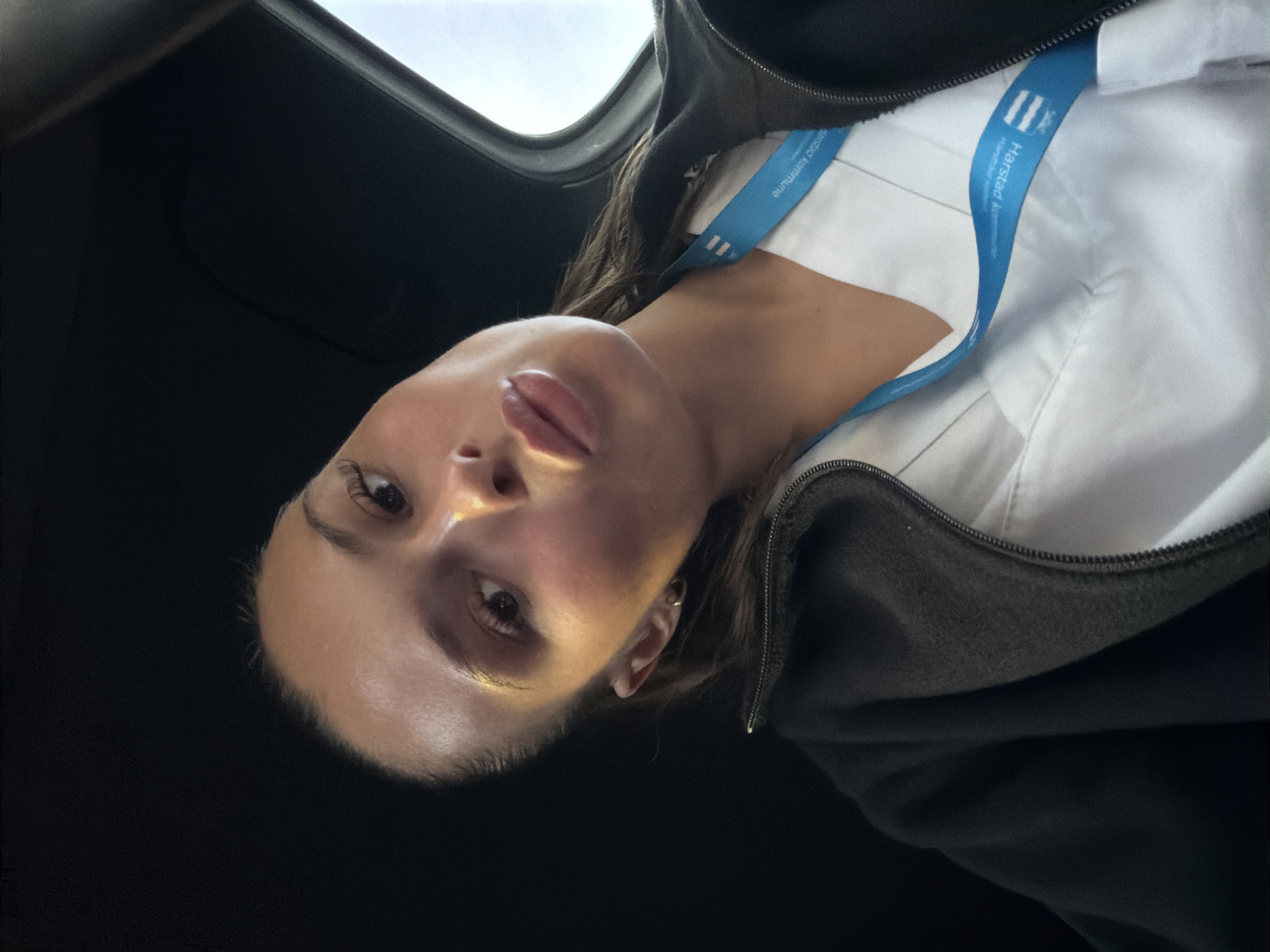 The image is a selfie of a young woman wearing a white uniform and a black jacket, with a blue lanyard around her neck. The lanyard has text that appears to read "Harstad Kommune," indicating the location is Harstad, Norway. The woman is in a car, with the interior of the car visible in the background, including the dark headliner and a portion of the window. The lighting suggests it is daytime. The woman is looking at the camera and has her lips pursed.