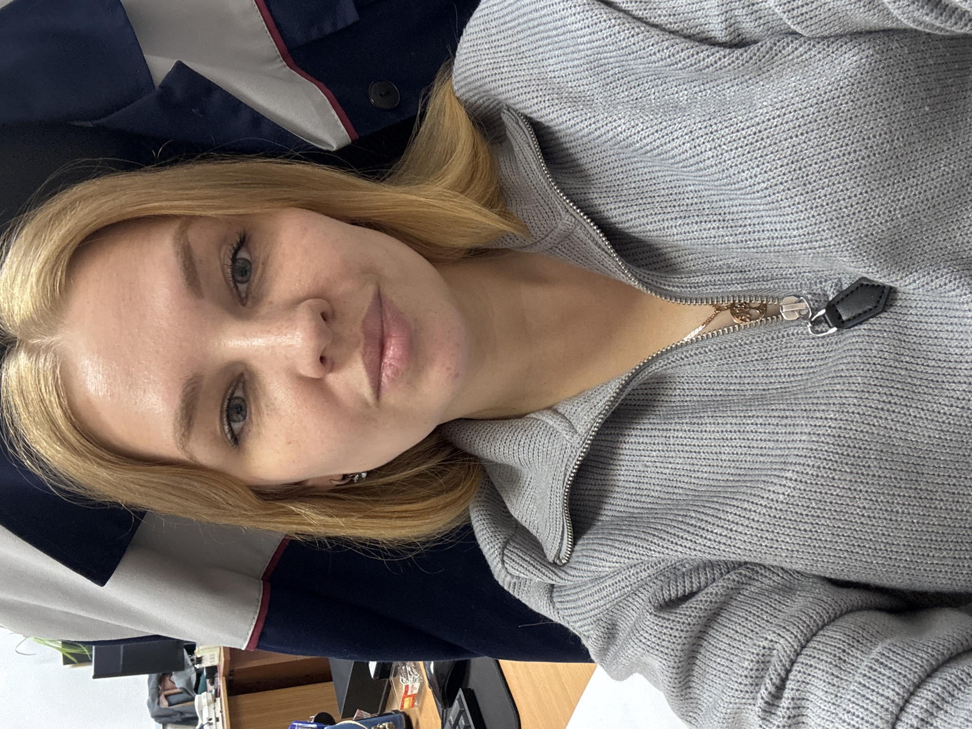 A young woman with light blonde hair and fair skin is posing for a selfie. She is wearing a gray knitted sweater with a partially zipped zipper. Behind her, there is a dark blue jacket with white and red accents. In the background, there is a desk with a computer monitor and some office supplies. The location is not identifiable as Rusanovo, Russia.