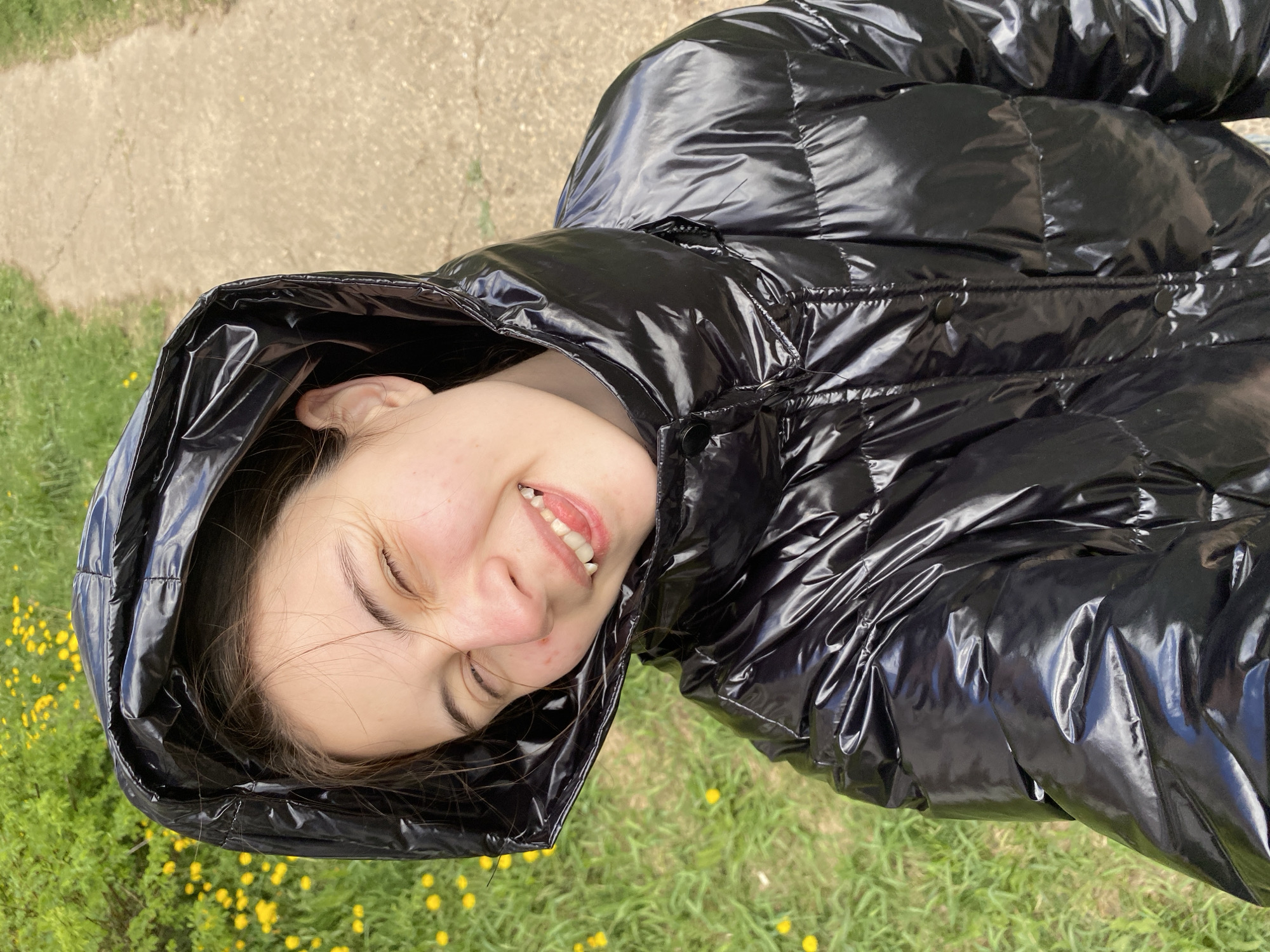 A young woman is lying on her back on grass dotted with yellow flowers. She is wearing a shiny black puffer jacket with the hood up, and she is smiling with her eyes mostly closed. In the background, there is a paved area with vegetation visible. The location cannot be verified as Votkinsk, Russia, based on the visual information available.