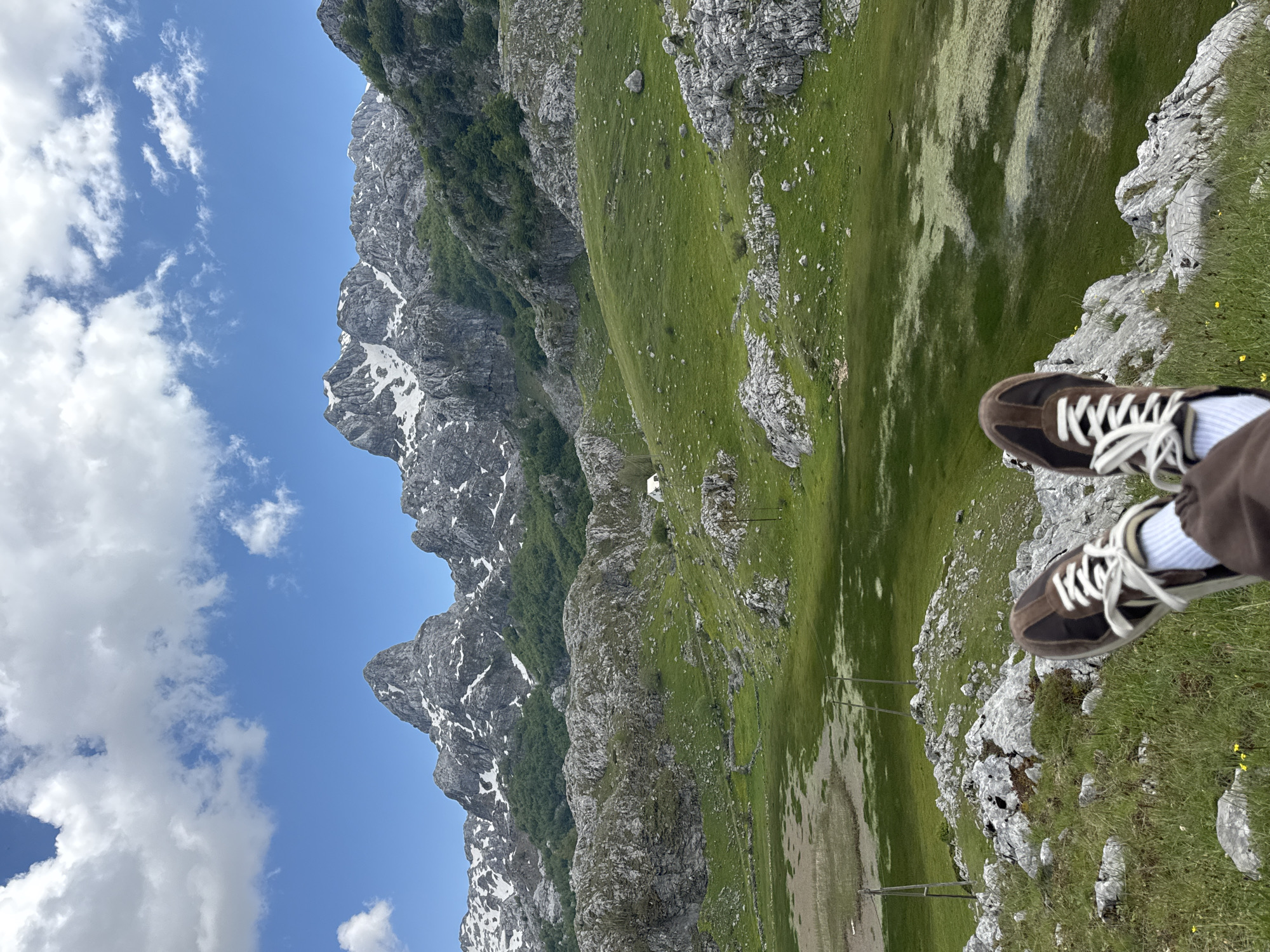 The image depicts a mountain landscape in Brskut, Montenegro. Jagged, rocky peaks with patches of snow are visible under a blue sky with scattered clouds. A green, grassy hillside slopes down towards a valley. In the foreground, a pair of brown shoes with white laces are visible, as if someone is sitting at the edge of a rocky outcrop overlooking the landscape. Power lines stretch across the valley floor.