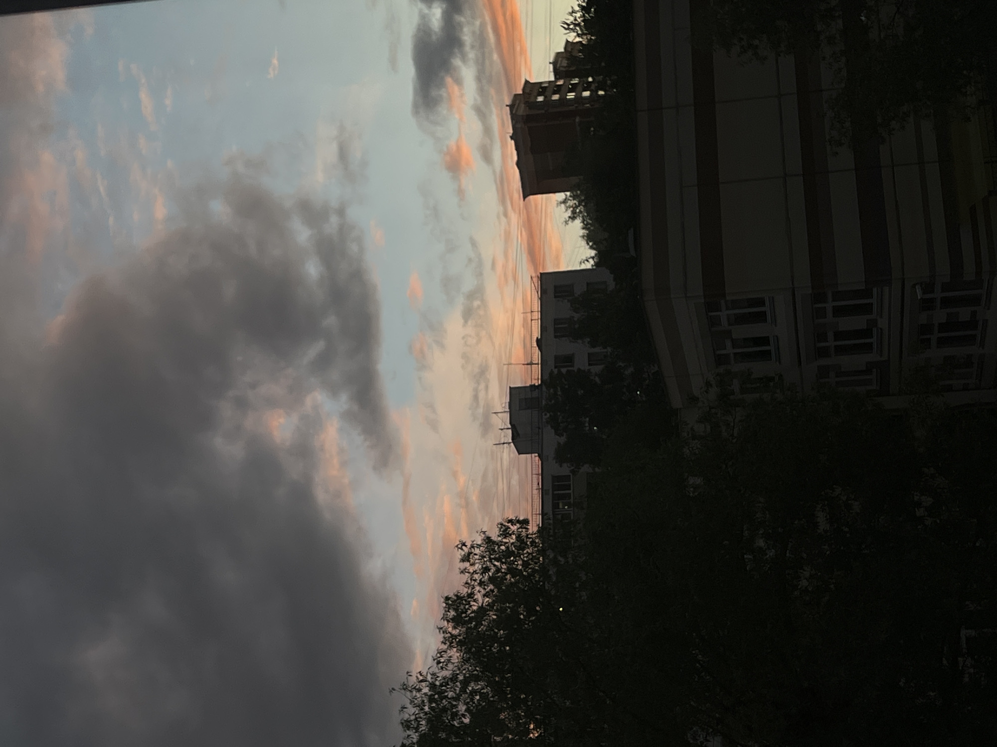 The image shows a view in Khimki, Russia at dusk. To the right are buildings, one with a striped facade, partially obscured by dark trees. To the left, the sky is visible, filled with dark clouds and some pinkish hues, indicating a setting sun. The image is rotated, giving a tilted perspective. No people are visible.