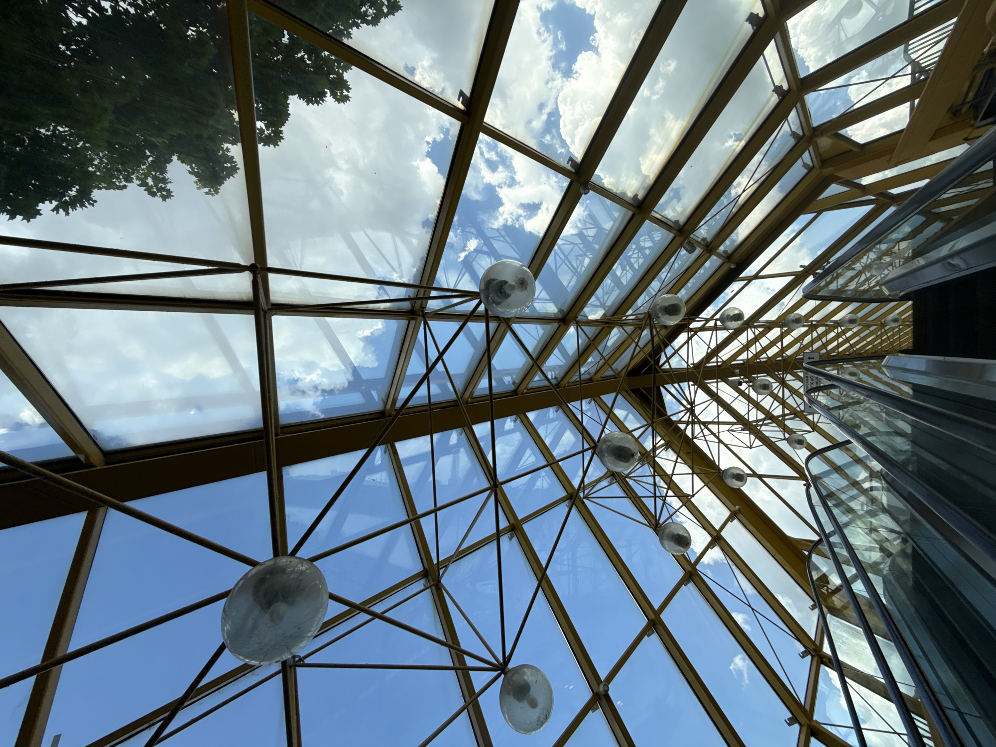 The image displays an upward-looking view of a large, geometrically structured glass ceiling, likely an atrium or skylight in a modern building. The framework supporting the glass panels is made of metal, appearing to be brass or a golden hue. Numerous spherical glass light fixtures are suspended at various points from this intricate framework.
Through the transparent glass panels, a bright blue sky with scattered white cumulus clouds is visible, indicating daytime. In the upper left section of the skylight, reflections of green tree foliage are evident on the glass surface. On the right side of the image, a sleek, vertically ascending structure, made of glass and dark panels, is partially visible. This structure appears to be part of an escalator or elevator system, reflecting the surrounding light and architectural elements. Faint, unreadable text, possibly in Cyrillic script, is discernable on one of the glass panels of this vertical structure.
The overall scene suggests a spacious, well-lit interior of a public building, possibly a shopping mall, airport, or hotel, designed to maximize natural light and offer a view of the sky. No specific activity or people are depicted in the static architectural shot.