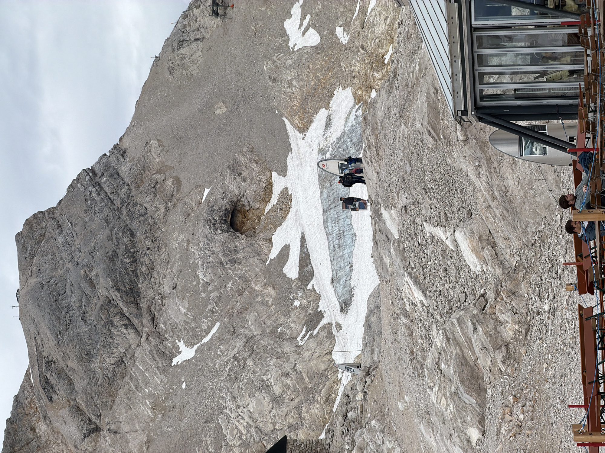 This image depicts a rocky mountainside with patches of snow and ice, likely at a high elevation. In the foreground, a modern building with a glass facade and a metal roof is visible, with outdoor seating areas consisting of wooden benches and tables. Two individuals are seated at one of these tables, facing away from the camera. Further up the slope, a group of four people are standing near a large, surfboard-shaped information display. Behind them, a portion of a glacier or large snowfield is visible, nestled within a rocky crevice. The background is dominated by the imposing, rugged mountain face, with evidence of weathering and rockfall. At the very top of the mountain in the background, a small structure, possibly a building or an observation point, can be seen. The sky is overcast and appears to be a light gray. The scene is suggestive of a tourist destination in a mountainous region. Given the context of Garmisch-Partenkirchen, Germany, this location is likely the Zugspitze, Germany's highest peak, known for its cable cars, ski resorts, and alpine scenery.