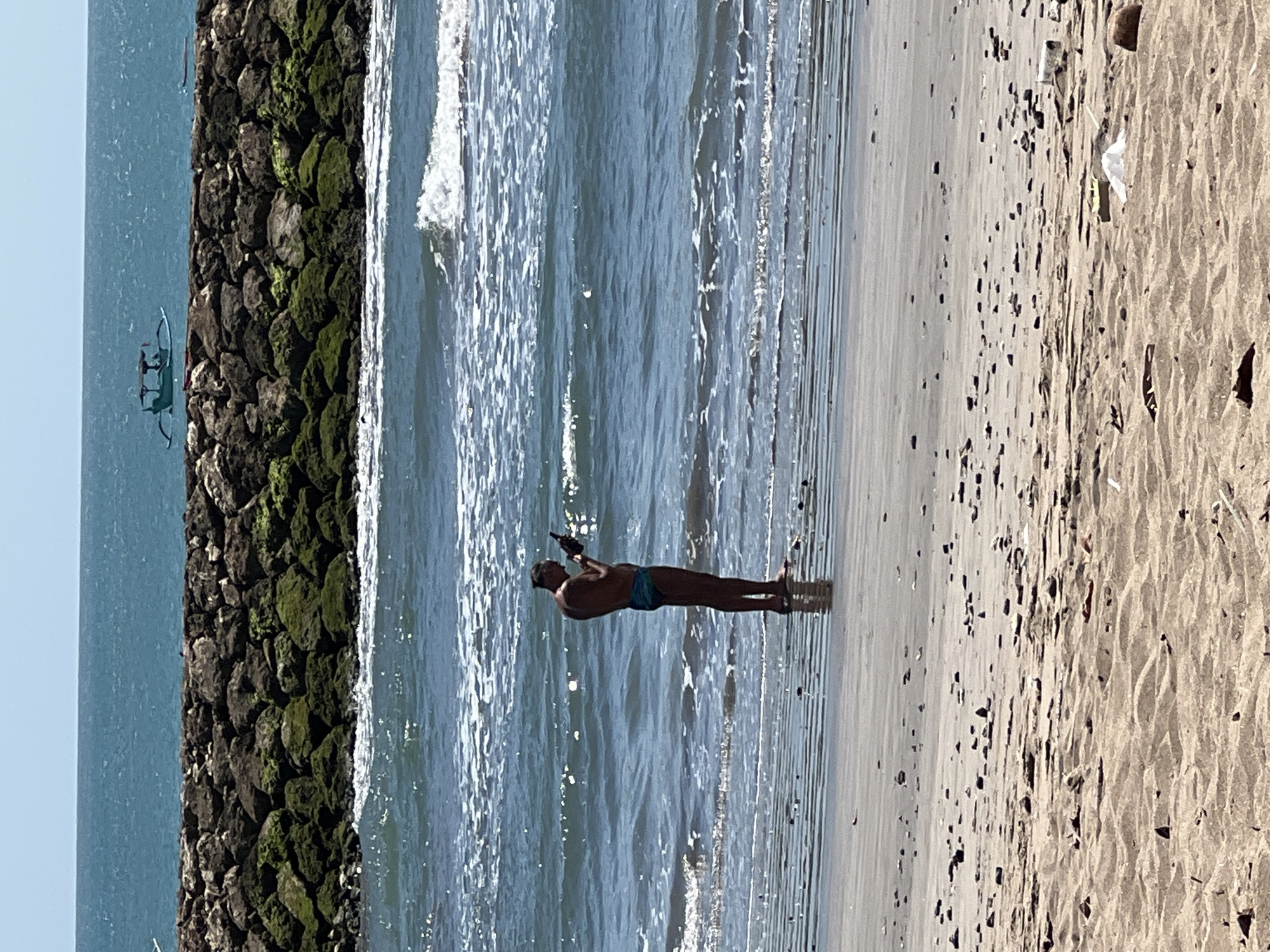 A lone man stands on the wet sand at the edge of the ocean in Kuta, Indonesia, looking at his phone. He is wearing blue patterned swim trunks. The ocean is calm with small waves breaking near the shore. A breakwater made of large, dark rocks, some covered in green moss, extends into the sea. In the distance, a small blue boat with a canopy is visible on the water. The sky is clear and blue. The foreground shows dry sand with small debris scattered across it, including rocks and some plastic waste.