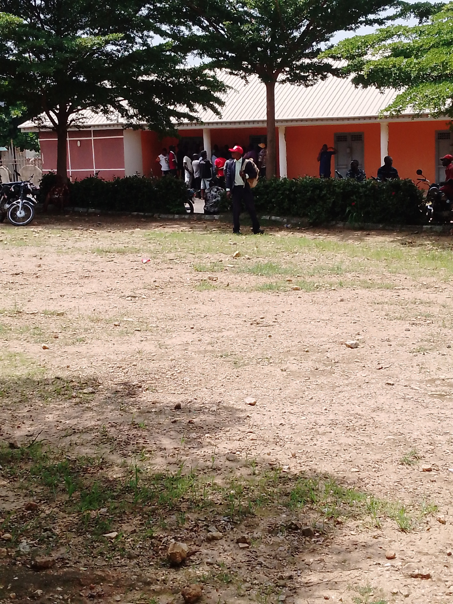 The image shows an outdoor scene in front of a building with an orange facade and a white, corrugated roof. Several people are gathered in front of the building, some appearing to be standing in line. A man in a dark suit and red cap is walking towards the viewer. To the left, there are two motorcycles parked. The ground is a mixture of dirt and gravel, with patches of grass. Large trees with green foliage are present in the foreground and midground, casting shadows on the ground. The scene appears to be a public gathering or an event taking place during daylight. The weather seems to be fair. There are no visible text elements in the image. The setting suggests a location in Akwanga, Nigeria, likely a public institution or a community center.