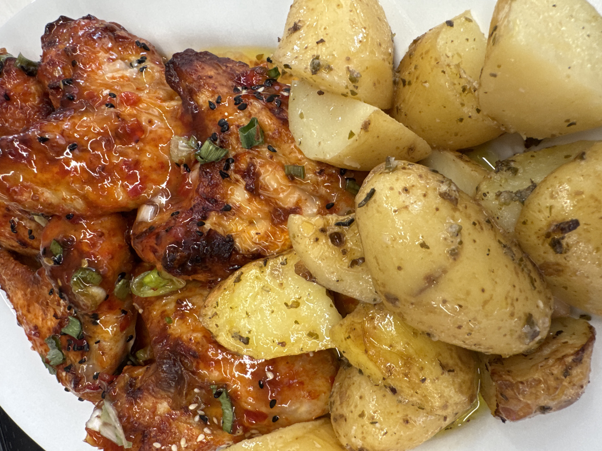 A close-up, overhead shot features a white plate filled with glazed chicken wings and roasted potatoes. The chicken wings are coated in a glistening, orange-red glaze and sprinkled with chopped green onions, white sesame seeds, and black sesame seeds. The roasted potatoes are small, golden-brown, and appear to be seasoned with herbs and oil, with some cut in half and others left whole. The lighting is bright, highlighting the textures and shine of the food. The background is a plain white surface, likely the plate itself. There are no people or recognizable landmarks visible, making it impossible to determine a location in Athens, Greece.