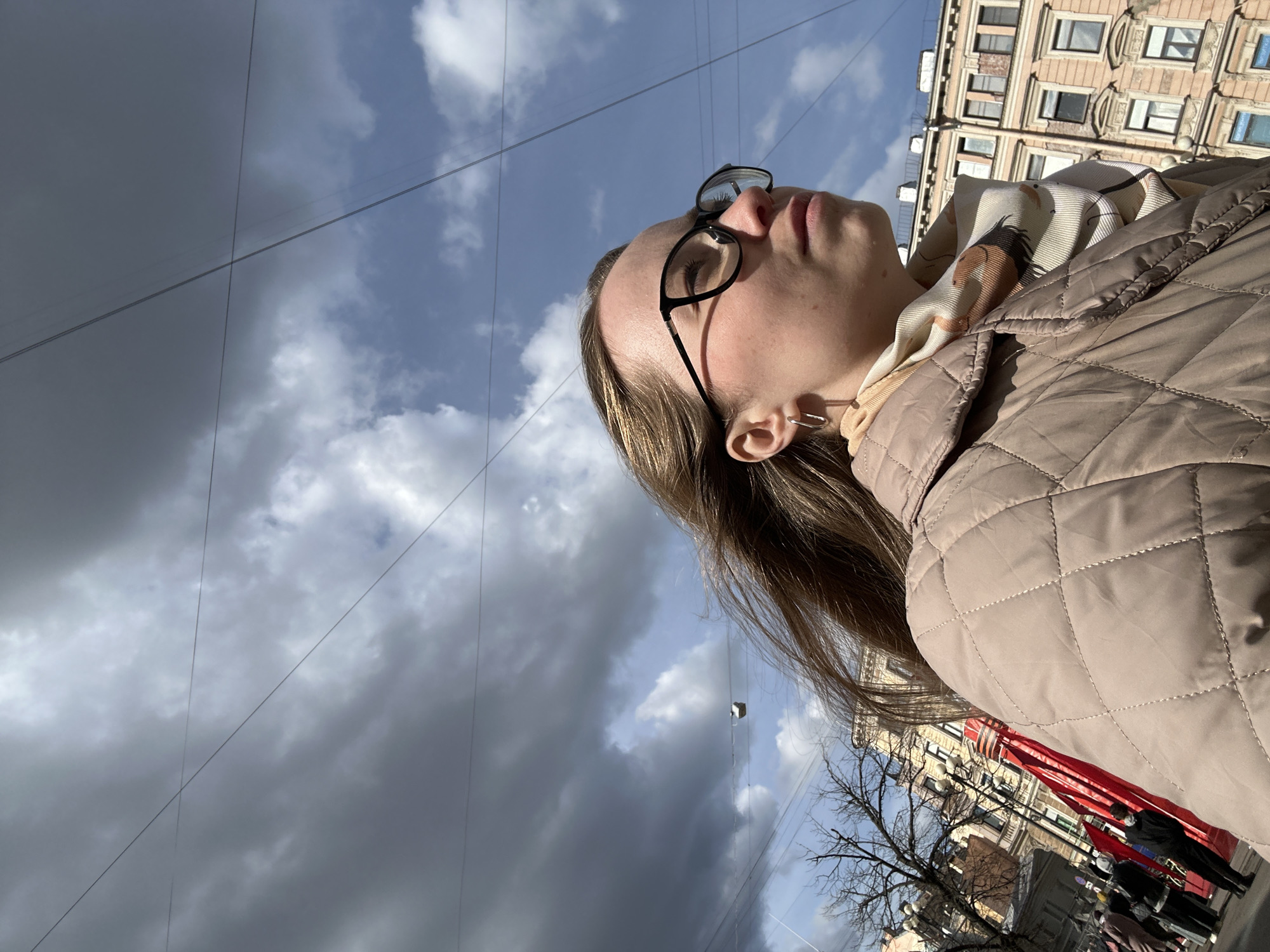 A low-angle outdoor shot captures a person, likely a woman, looking upwards with a contemplative expression. She is seen in profile from the chest up, wearing a light beige quilted jacket and a patterned scarf featuring an equestrian-like design around her neck. Black-framed glasses rest on her nose, and a silver ear cuff is visible on her left ear. Her light brown hair falls down her back.
The background is dominated by a dramatic sky, split between patches of clear blue with white clouds and areas of dark, heavy grey clouds, suggesting dynamic or changing weather. Numerous thin overhead wires crisscross the sky, adding an urban element. To the right, blurred in the background, stands a multi-story building with a classic European architectural style, featuring numerous windows. Below this building and partially obscured by a bare-branched tree, faint red banners or decorations are visible, suggesting a civic display or event. The strong sunlight casts shadows on the person's face and jacket, indicating it is daytime. No visible text is present in the image.