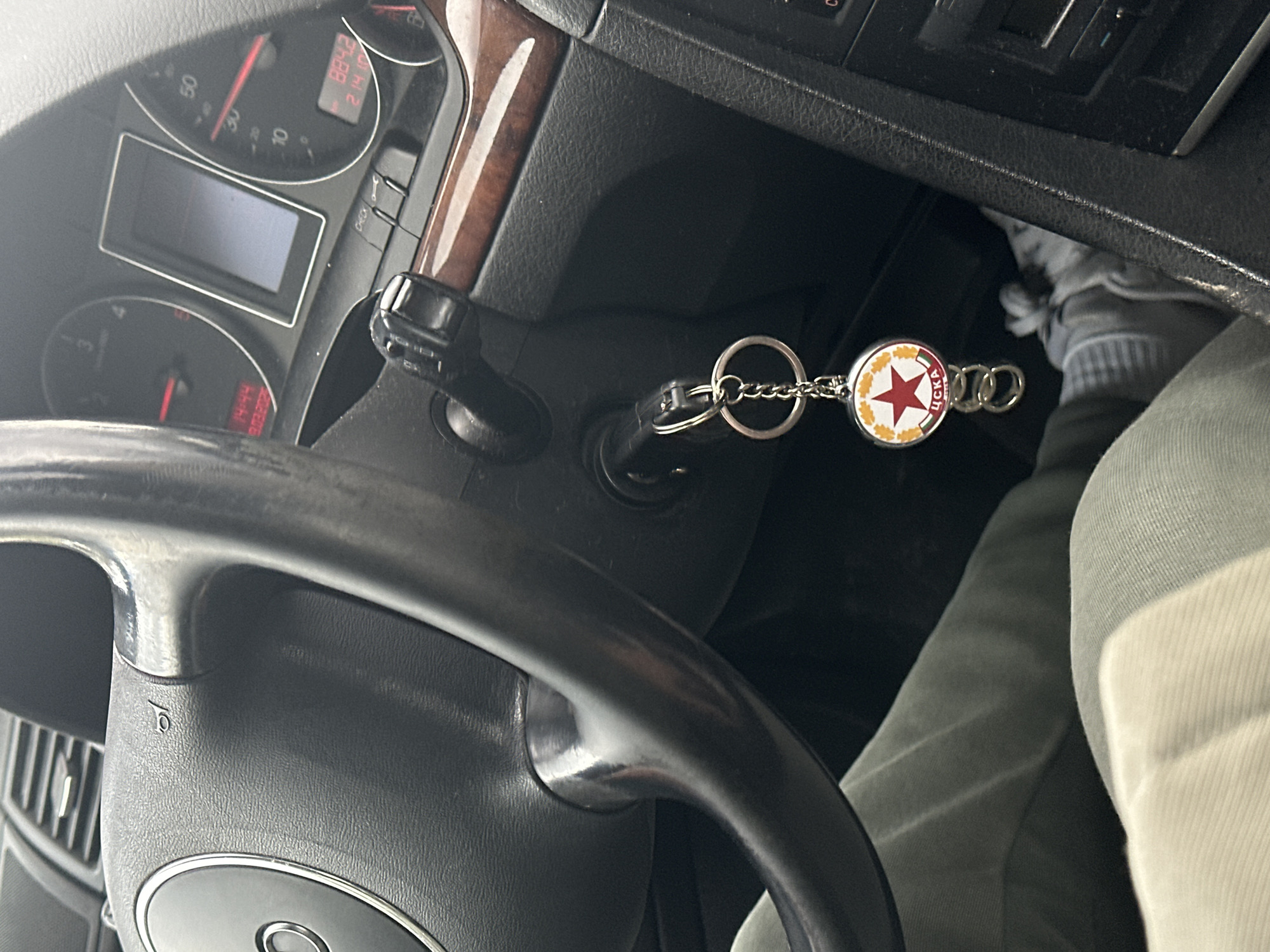 The image depicts the interior of a stationary Audi vehicle from the driver's perspective. A black three-spoke steering wheel with a central Audi logo is on the left. The instrument cluster displays the time "14:44" and date "19.03.2023" on the left digital screen, and an odometer reading "884.2" and "214.0" on the right. A key is inserted into the ignition, from which a silver metal keychain is suspended. The keychain features a circular medallion with a prominent red five-pointed star, encircled by golden laurel leaves on a white background, and the Cyrillic inscription "ЦСКА" (CSKA) on a red band beneath the star. Three small, interlinked silver rings are attached below the medallion. The lower section of a person's left leg, clothed in light grey trousers, is visible, along with the right foot wearing a white sock and a grey athletic shoe near the floorboard. The "ЦСКА" (CSKA) emblem on the keychain identifies the scene's location as Sofia, Bulgaria, home of the CSKA Sofia football club.