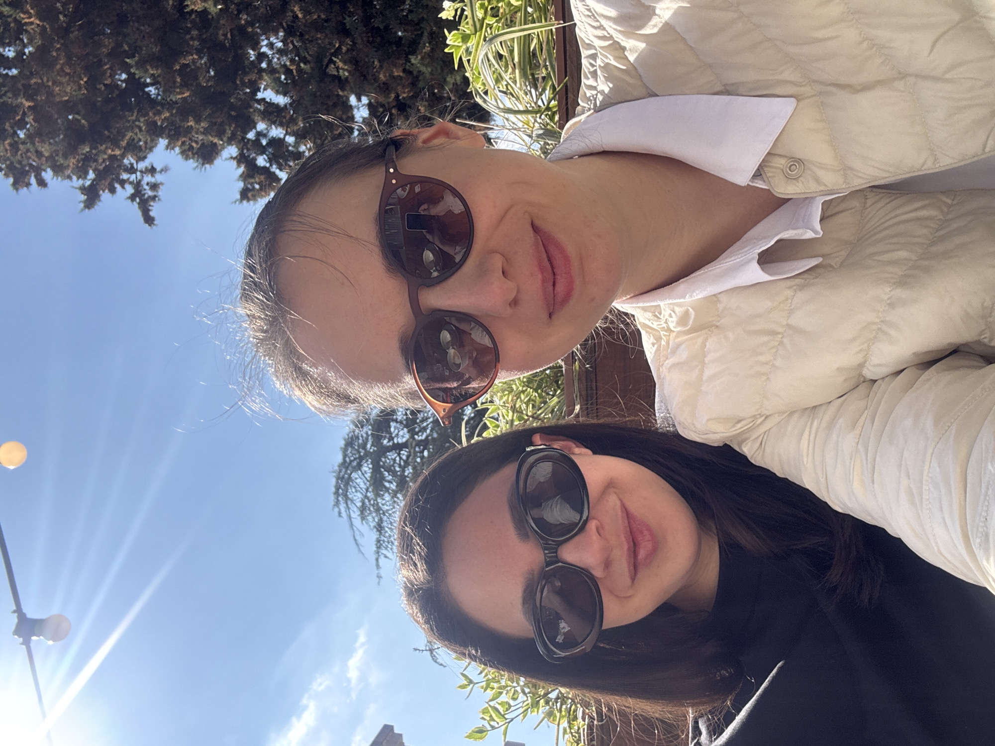 Two women are taking a selfie outdoors in Tbilisi, Georgia. The woman on the top is wearing a light-colored jacket and brown-rimmed sunglasses. The woman on the bottom is wearing a black top and dark-rimmed sunglasses. Trees and sky are visible in the background. Street lighting is visible in the background. The weather appears to be sunny.