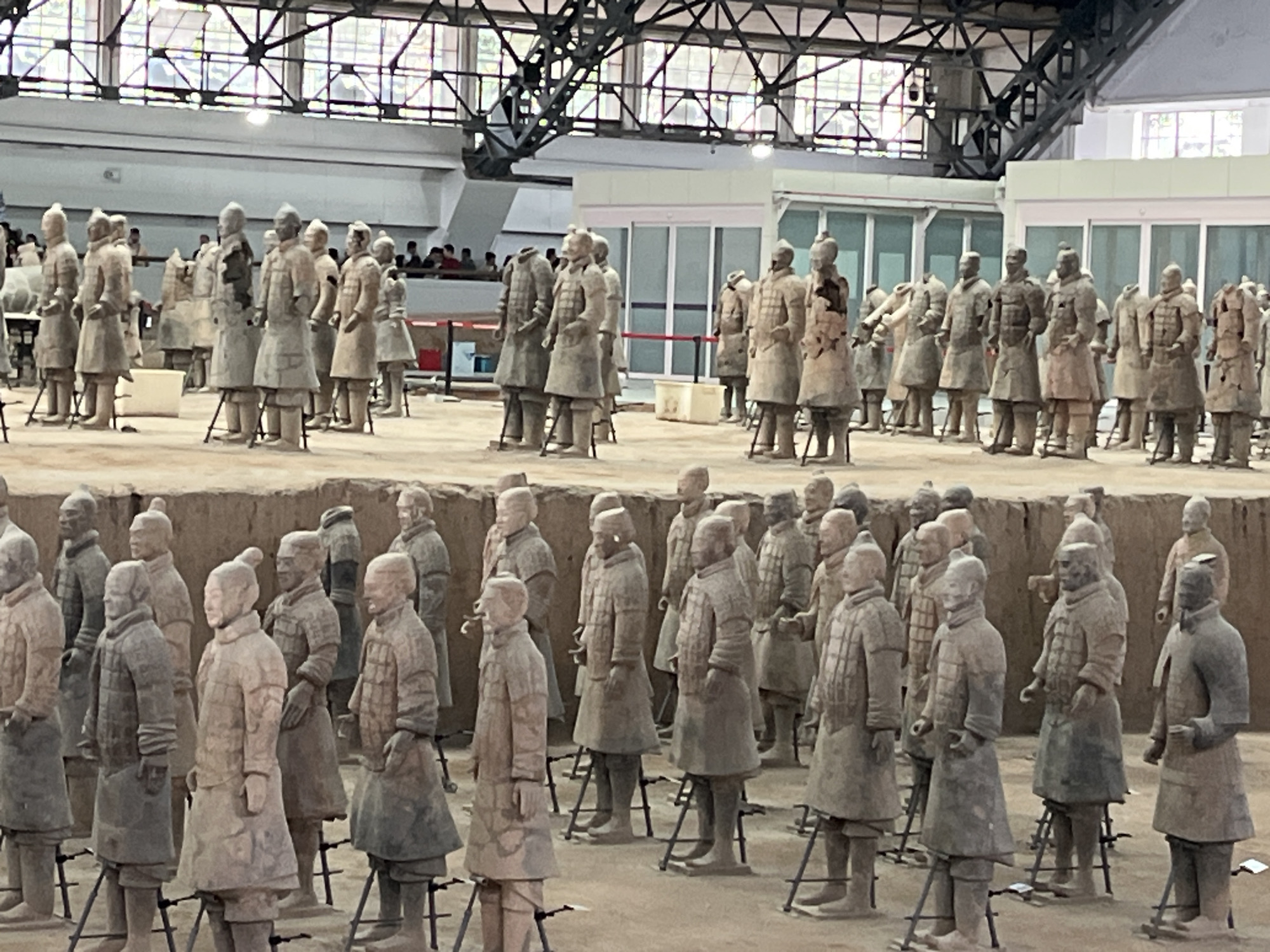 This media file depicts the Terracotta Army, a collection of terracotta sculptures depicting the armies of Qin Shi Huang, the first Emperor of China. The subjects are numerous life-sized terracotta figures of soldiers, chariots, and horses, arranged in battle formations within a large, excavated pit. The soldiers are depicted in various ranks and poses, wearing armor and carrying weapons, though many of these are no longer present.
The scene is set within a large, open-air museum or archaeological site, indicated by the excavated earth walls and the visible structure of a building with a metal truss roof and large windows overhead. The lighting suggests daytime, with the overall atmosphere being one of historical significance and preservation. There are no explicit indicators of weather conditions.
Notable details include the immense scale of the display, with rows upon rows of figures stretching into the distance. The figures themselves show individualistic features, suggesting a high level of craftsmanship. The presence of people in the background, observing the display, hints at the site being a tourist attraction. There is no visible text in the media file. The overall impression is one of awe and wonder at the historical and artistic achievement represented by the Terracotta Army.