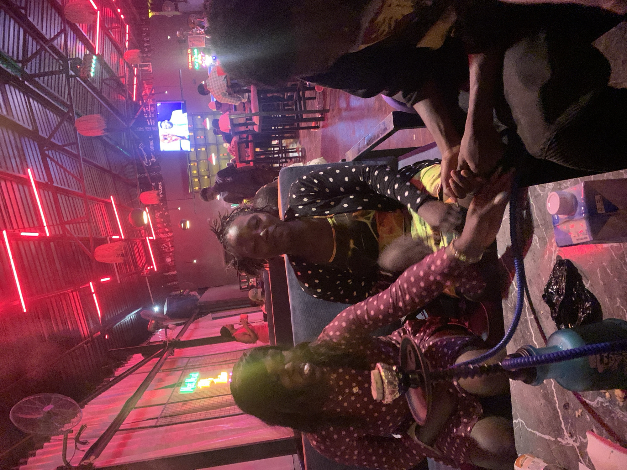 The image depicts a group of people socializing at a hookah lounge on Rukuba Road, Nigeria. In the foreground, three individuals are seated at a marble-top table, engaged in conversation and activity. A woman on the left, wearing a polka-dot dress, holds a hookah pipe. To her right, another woman in a polka-dot top and patterned dress is visible. Partially obscuring the view is a third individual on the far right. The background reveals a lively ambiance with red neon lighting illuminating the ceiling and walls, a large fan mounted on the wall, and other patrons seated at tables and a bar area. A television screen displaying an image of a man is mounted high on the wall. The overall scene suggests a casual and vibrant nighttime social gathering.