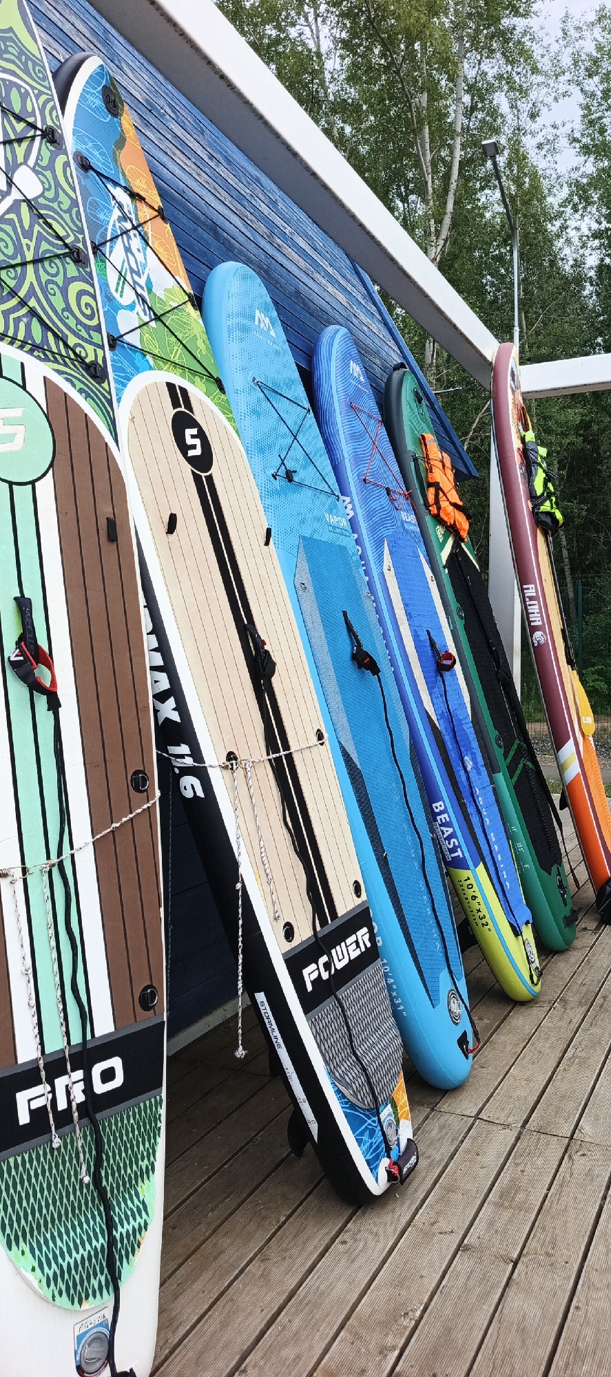 The image shows a collection of stand-up paddleboards (SUPs) leaning against a blue wooden wall. The SUPs are of various colors and designs, with some featuring prominent brand names and specifications such as "MAX 11.6", "BEAST 10'6\"x32\"", and "PRO". Several SUPs have ropes attached, and some have orange life vests draped over them. The boards are arranged in a line, with the closest one on the left being white with green, brown, and white stripes. The next board is tan with a black stripe and a black circle containing the number "5". The remaining boards are predominantly blue, green, and maroon. The scene appears to be outdoors, with trees visible in the background and a wooden deck in the foreground. The lighting suggests it could be daytime. The overall impression is one of readiness for water activities, with the SUPs neatly stored. No people are visible in the image.