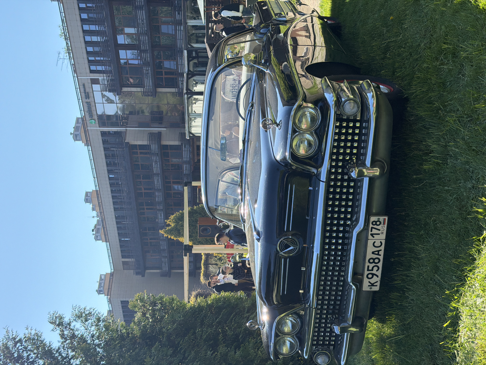 The image depicts a black classic car parked on a grassy area, possibly in Repino, Russia, as indicated by the Russian license plate "K958AC 178 RUS". The car is facing the camera, showcasing its chrome grill and headlights. Behind the car, there is a building with a modern architectural style, featuring multiple windows and balconies. In the background, there is a group of people standing and talking. The sky is clear and blue. The general setting appears to be a car show or exhibit.