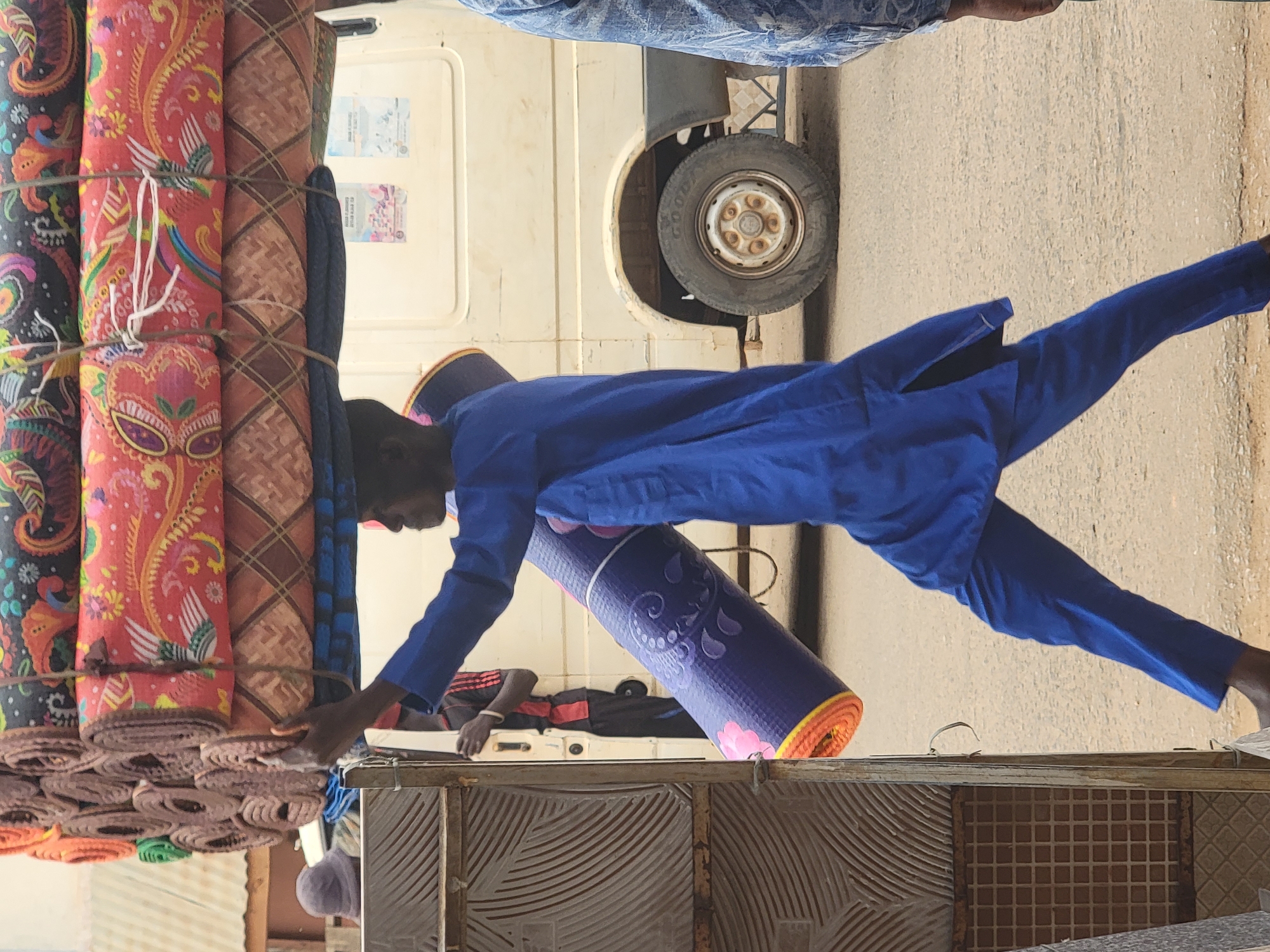 In Maiburiji, Nigeria, a man in a blue outfit is standing beside a cart laden with rolled carpets. He is handing a rolled purple and orange yoga mat to someone lying on the cart. The cart is positioned next to a white vehicle. The area appears to be a street.