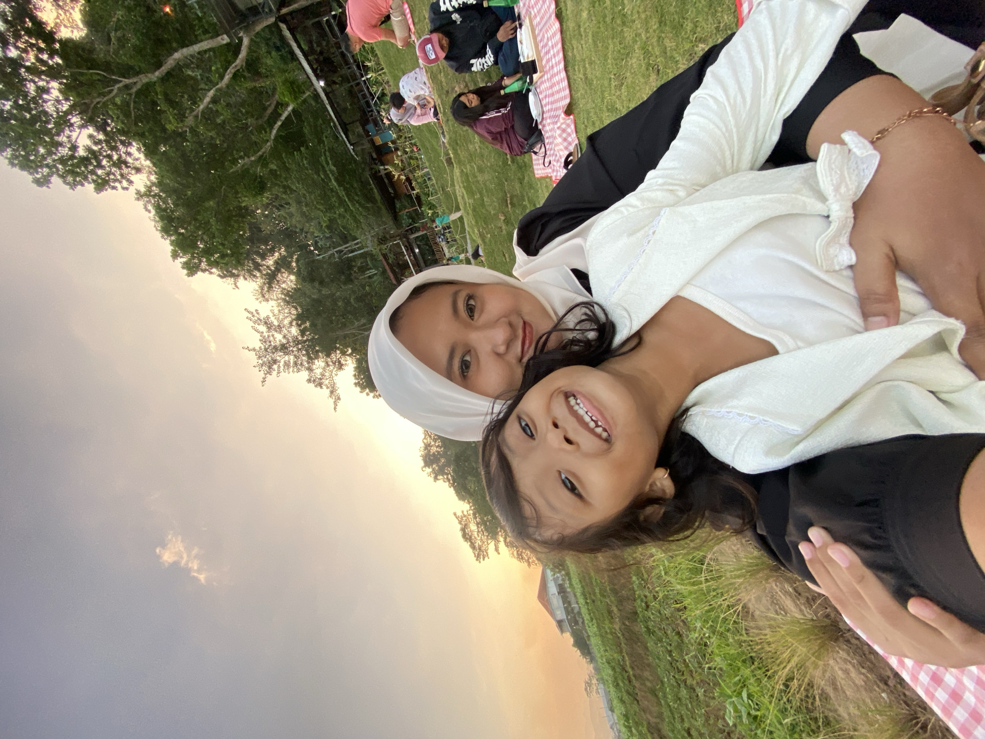 In the city of Kesiman, Indonesia, at sunset, a woman in a white hijab and a young girl with dark hair are in the foreground, taking a selfie. The woman is wearing a black outfit with a white blouse underneath, while the girl is wearing a white jacket over a white top. They are sitting on a red and white checkered picnic blanket on a grassy hill. In the background, other people are seen picnicking on blankets on the lawn. Trees and some structures are visible in the distance. The sky is a soft orange and yellow, indicating the setting sun.