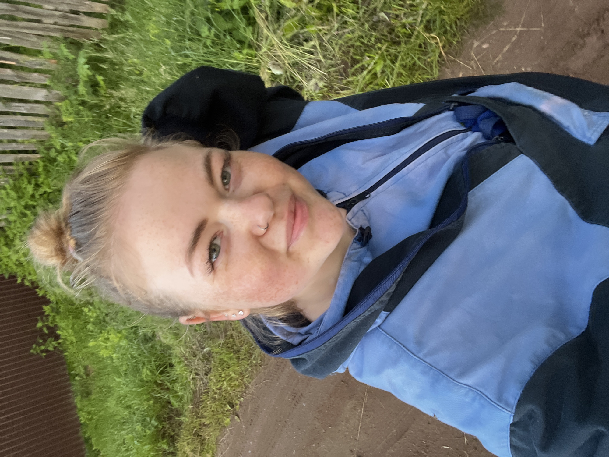 A young woman with blonde hair pulled up in a bun is lying down, taking a selfie. She has light skin with freckles across her nose and cheeks, and green eyes. She is wearing a light blue and black jacket. The background is an outdoor scene with green grass, a dirt path, and a wooden fence. The lighting suggests it is daytime, and the lush green foliage implies warm weather. The woman has a relaxed and happy expression, smiling gently at the camera. She is wearing a nose ring and earrings. The context of Ольховка, Russia is provided for location. There is no visible text in the image.
