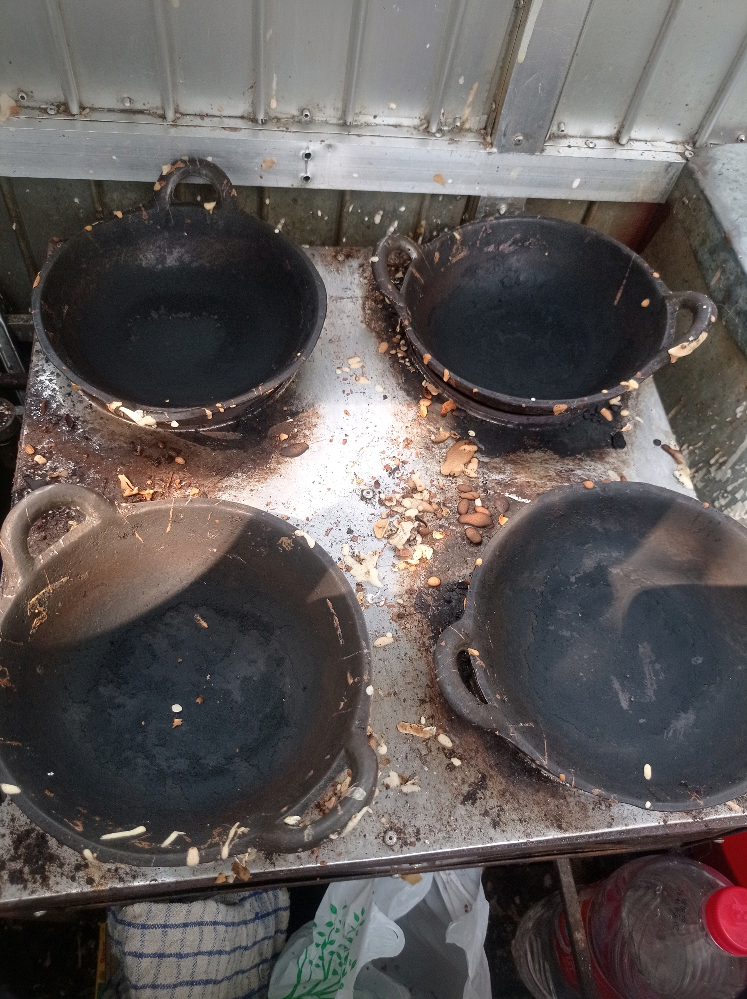A top-down, medium shot captures four dark, cast-iron pans resting on a metallic surface. The pans are round with two handles each and appear to be used for cooking. The surface beneath the pans is dirty and scattered with food debris, including what appear to be peanut shells and small pieces of food. In the background, a corrugated metal surface is visible, with some white residue splattered on it. To the bottom of the image, partially visible, are a white plastic bag with a green leaf pattern, a rolled-up checkered cloth, and a clear plastic bottle with a red cap, containing a reddish liquid, likely a soft drink. The scene is likely from a street food stall or a small eatery in Tuban, Indonesia, given the type of cookware and the general setting.