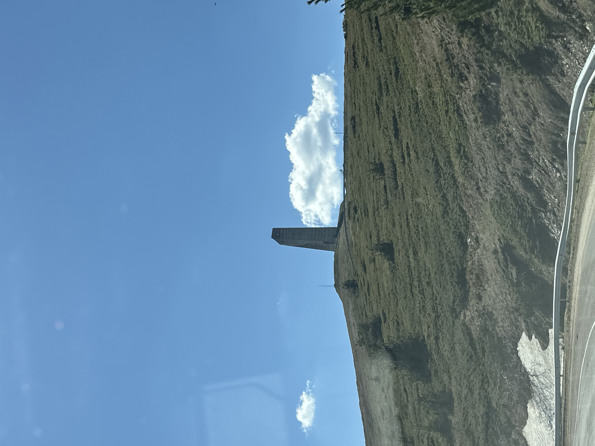 The image shows a concrete tower on the edge of a cliff face in Beli Osam, Bulgaria. The cliff is covered in vegetation. A road runs along the base of the cliff. There are clouds in the blue sky above the tower. The image was likely taken from a moving vehicle.