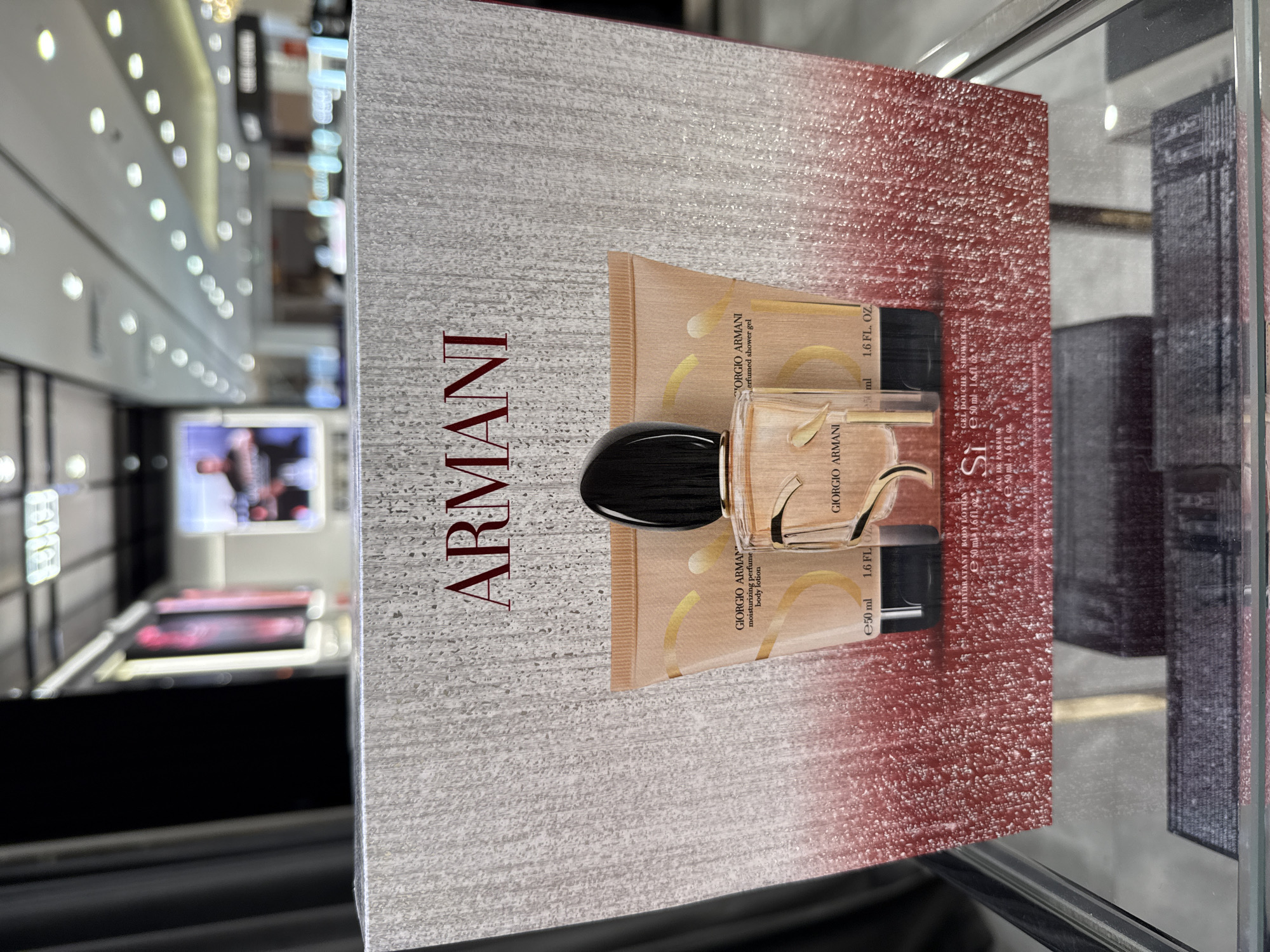 A Giorgio Armani Si perfume gift set is displayed on a shelf in a retail environment. The set includes a perfume bottle and two tubes, likely a body lotion and shower gel, all featuring the "Si" branding. The packaging is predominantly silver with a gradient of red at the bottom. The background shows a blurred view of a shopping mall, with display screens and ceiling lights visible, suggesting an indoor commercial setting.