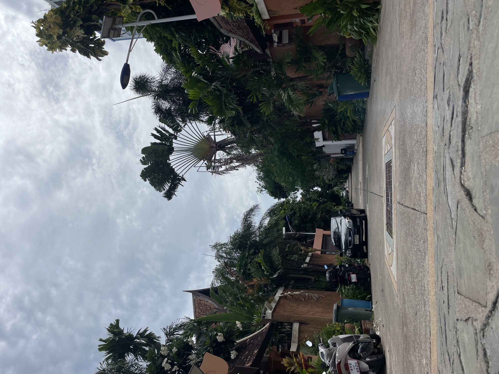 The image captures a view looking down a quiet street in Pattaya City, Thailand, with overcast skies. The street is paved with concrete and features a visible drainage grate in the center. On either side of the street, there are residential buildings with reddish-brown tiled roofs and peach-colored walls. Lush tropical vegetation, including palm trees and flowering plants, lines the street and surrounds the houses. Parked on the left side of the street are two scooters, one white and one black, and a black sedan is parked further down on the right. Two green trash bins are visible next to the scooter on the left. The overall scene depicts a residential area in a tropical climate.