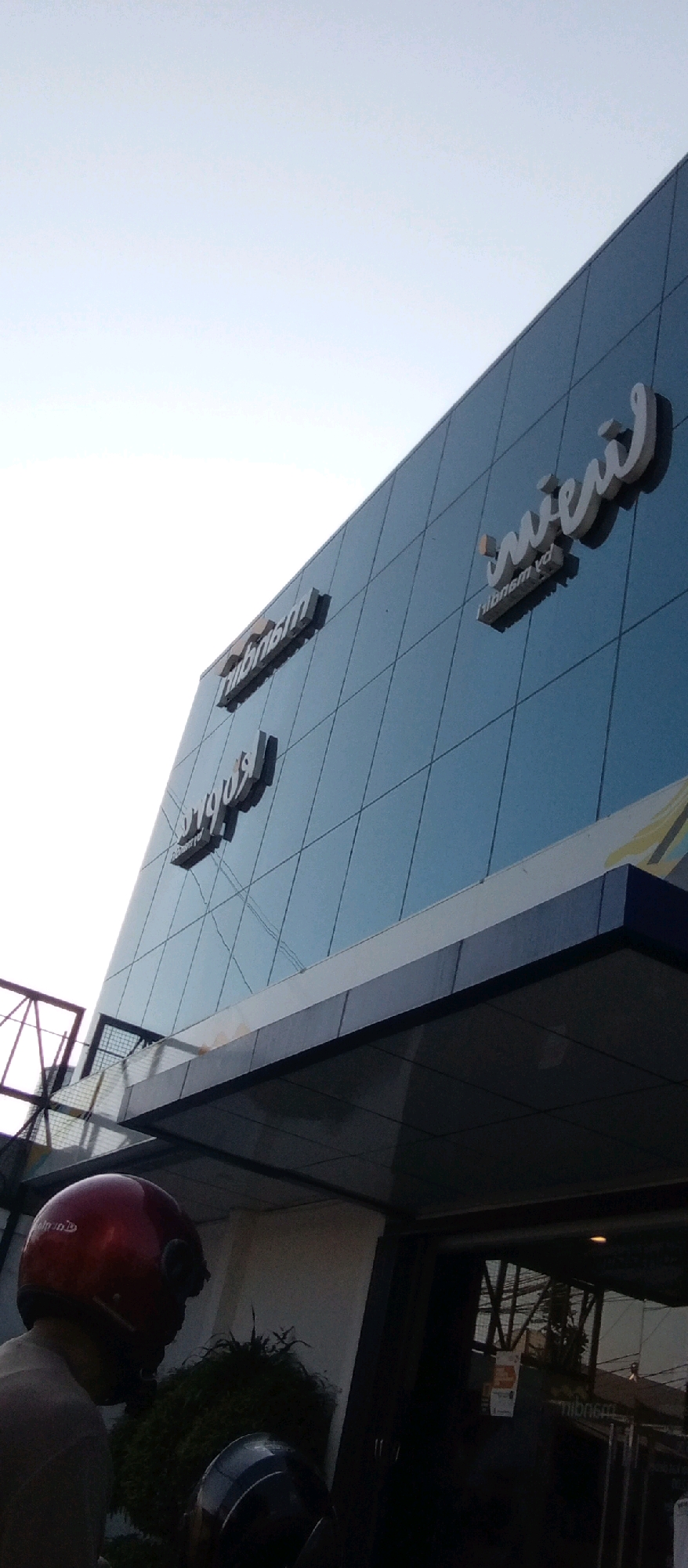 This image captures an exterior view of a modern building with a reflective blue glass facade, likely located in the city of Kediri, Indonesia. The building features prominent, raised signage in white, stylized lettering. One sign reads "Tribun" and another appears to be "Indi" or "Indri" followed by what looks like "am". Below the "Tribun" sign, there is another sign, possibly a reflection, that reads "ribunam yd". The building has a sleek, contemporary design, with a darker band running along the top of the entrance area. In the foreground, a person wearing a red helmet is partially visible, seated on a scooter, facing away from the camera and towards the building. Another dark-colored helmet is visible on the seat of a scooter in front of the person. The environment suggests a daytime setting with bright, overcast sky. The overall impression is one of a commercial or institutional building.