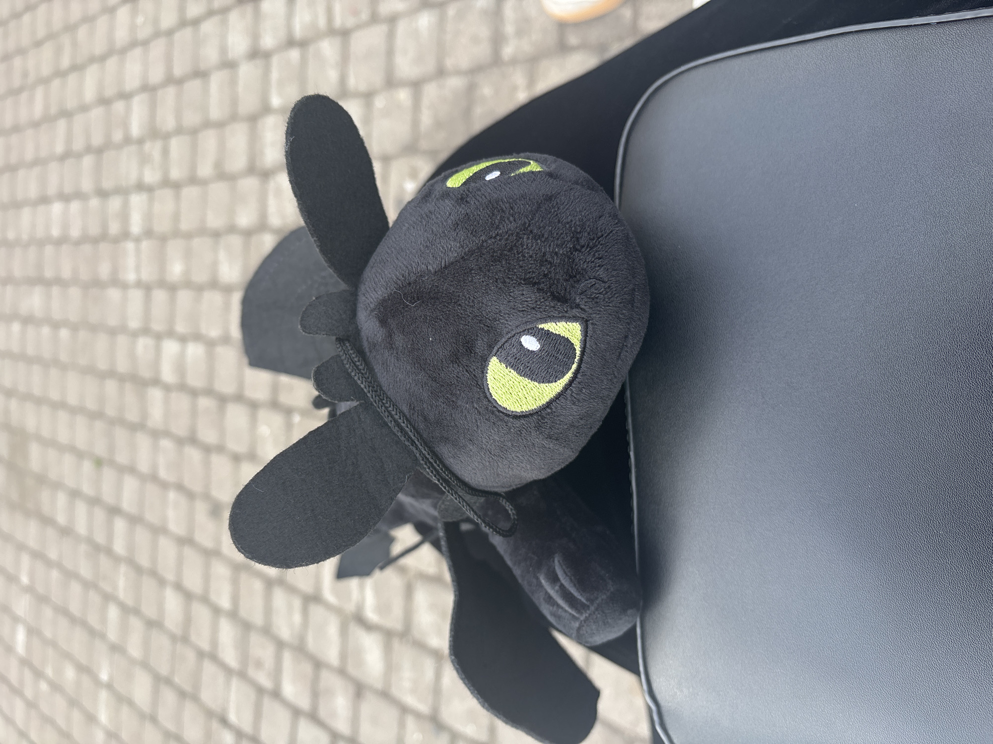 A black plush toy resembling the character Toothless from "How to Train Your Dragon" is placed on top of a dark-colored bag. The toy is positioned with its head facing the viewer, its green eyes with white pupils clearly visible. Its black wings are spread, and its body is slightly angled. The background consists of a light-colored, textured surface that appears to be pavement made of interlocking rectangular tiles. The lighting suggests an outdoor environment, possibly during the daytime, with the sun casting some shadows. The scene is cropped to focus primarily on the toy and the bag it rests on. The location is identified as an unknown area within the city of Unknown, Russia.