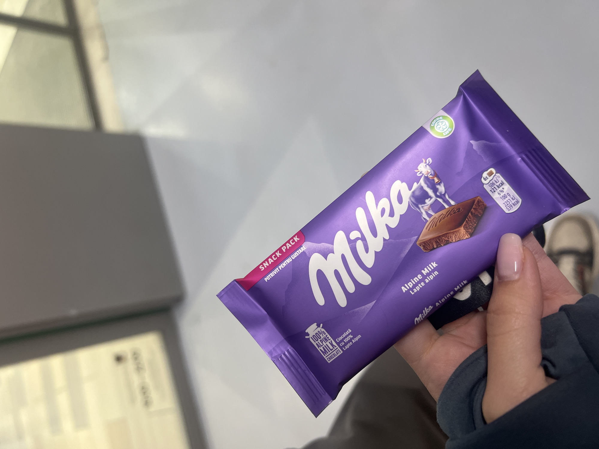 A person is holding a purple Milka Alpine Milk snack pack in their hand, with the packaging clearly visible, displaying the brand name, a cow graphic, and nutrition information. The background includes a blurred grey surface, possibly part of an interior, and a glimpse of a shoe. The text on the packaging is in Romanian and English. The photograph appears to have been taken indoors with natural light. The location is Sofia, Bulgaria.